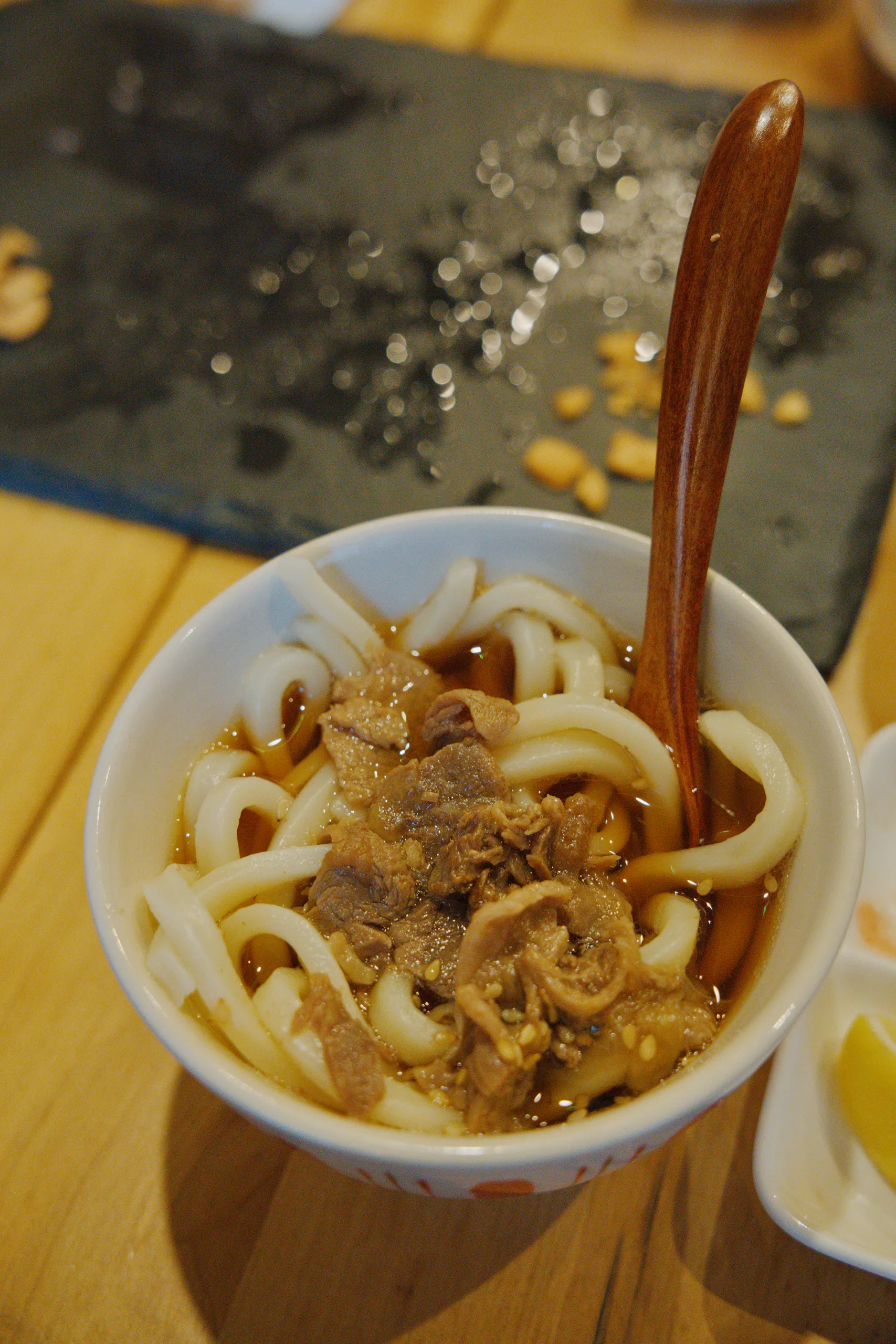 Bowl of curled noodles topped with meat and garnish, wooden chopsticks laid across the rim on a yellow surface.