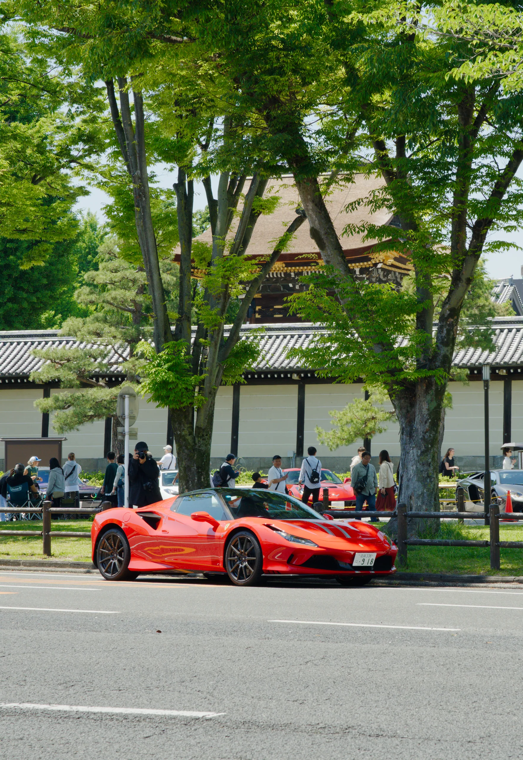 A red Ferrari parked on a street in front of a park pavilion with people dining in the background.