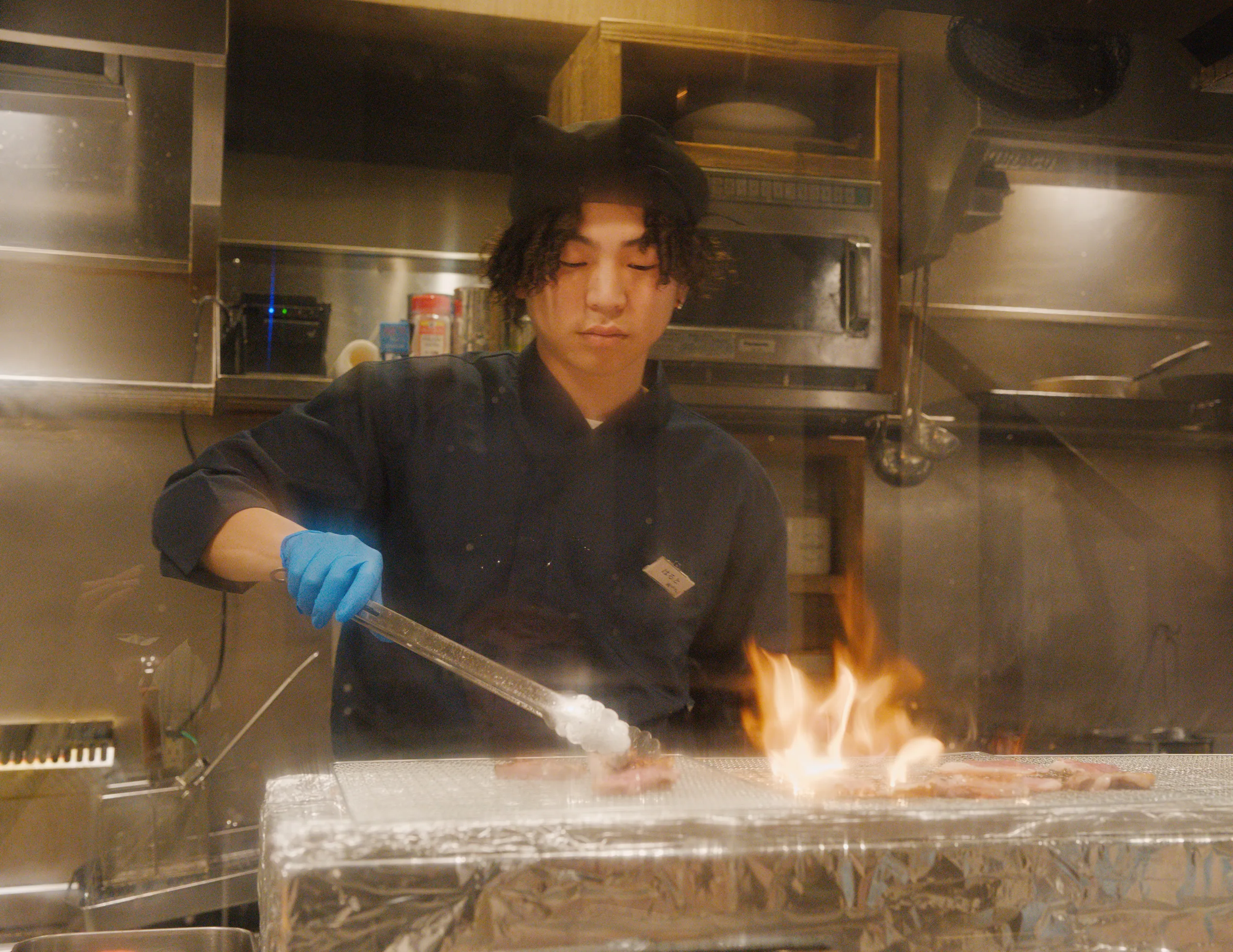 A chef in blue gloves tends flames at a cooking counter, viewed through a glass partition.