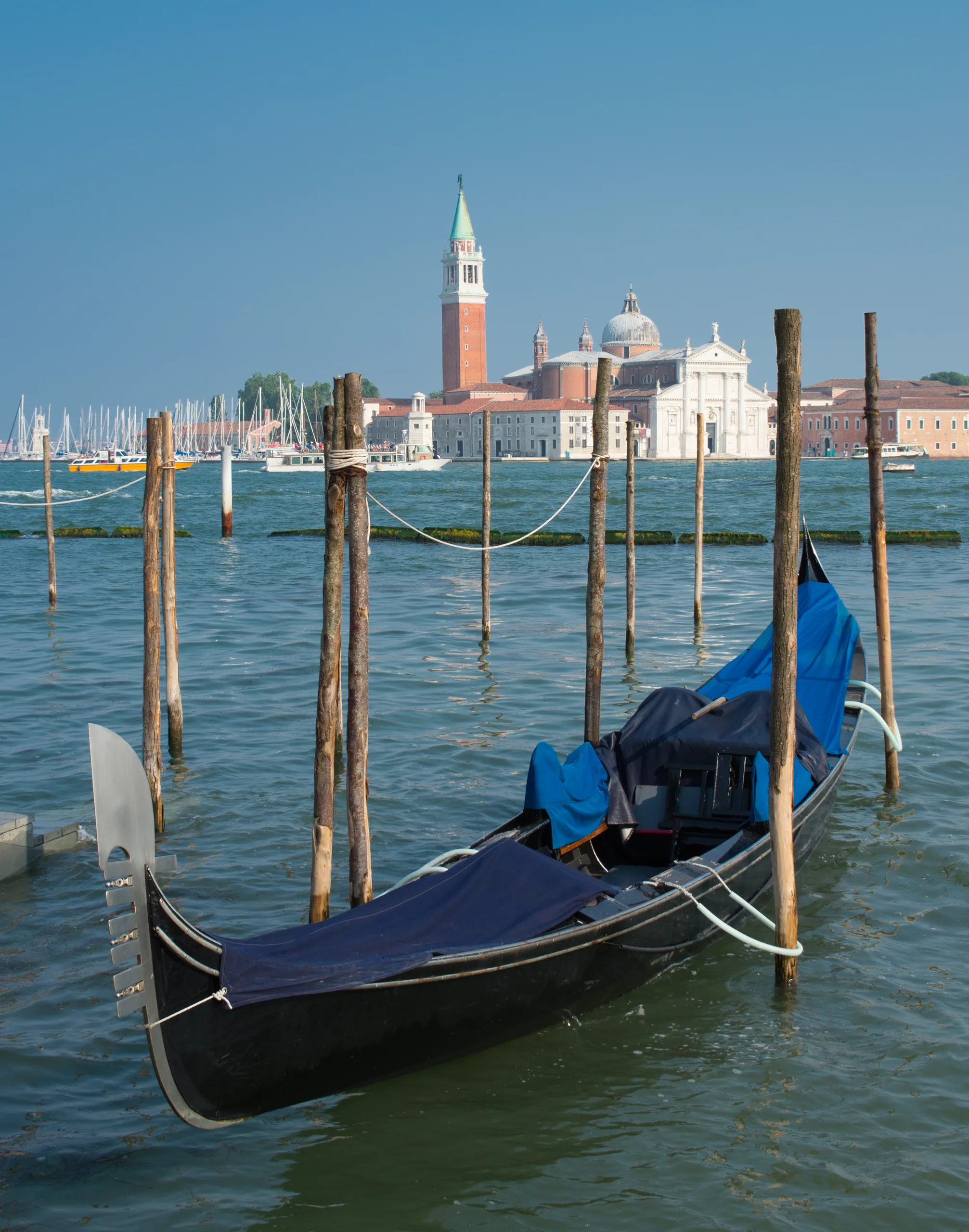 A moored gondola under its blue cover beside striped mooring poles, San Giorgio Maggiore across the water behind.