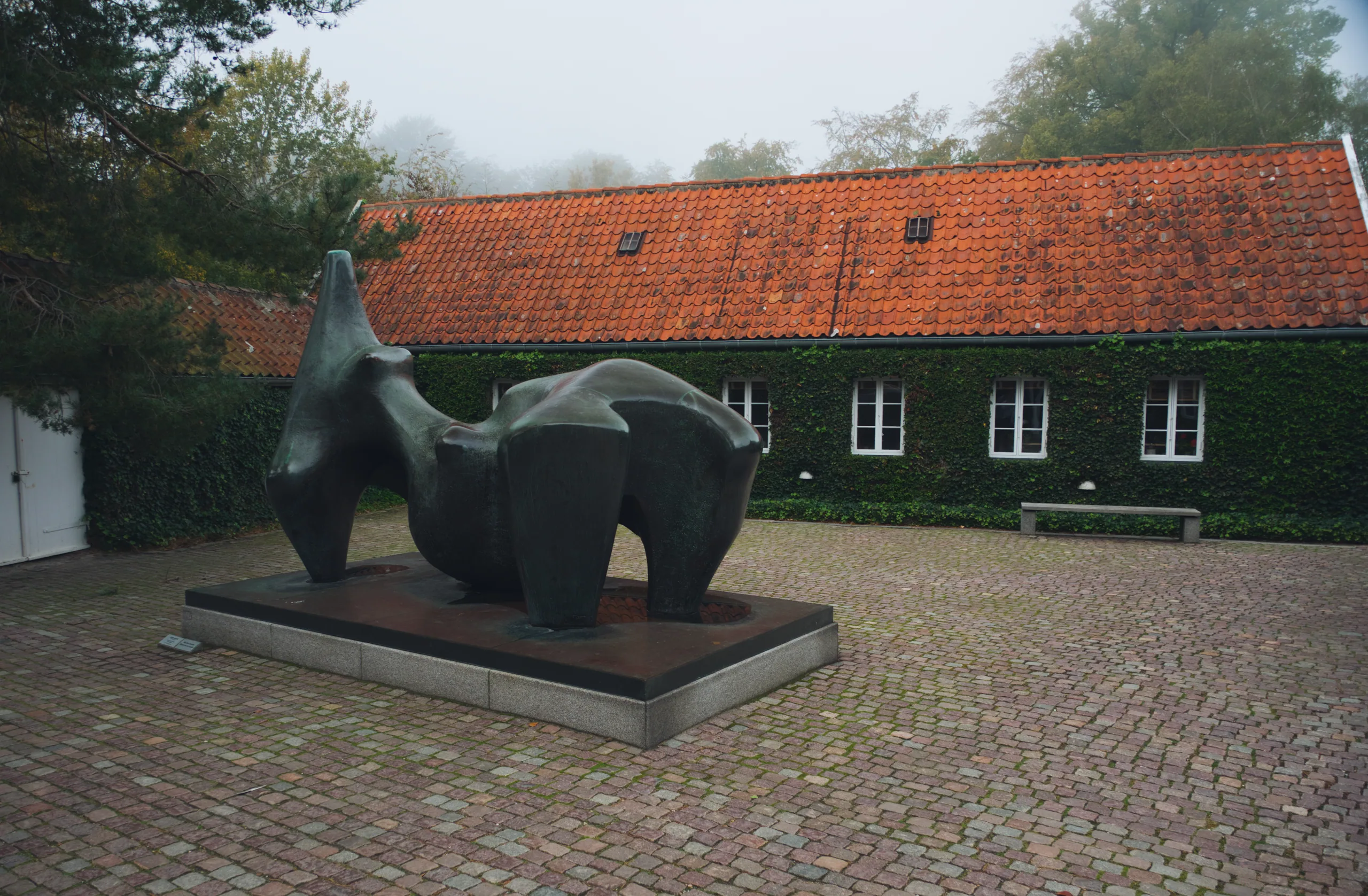 An abstract bronze sculpture of rounded interlocking forms on a stone plinth in a cobbled courtyard, an ivy-covered building with a red tile roof behind.