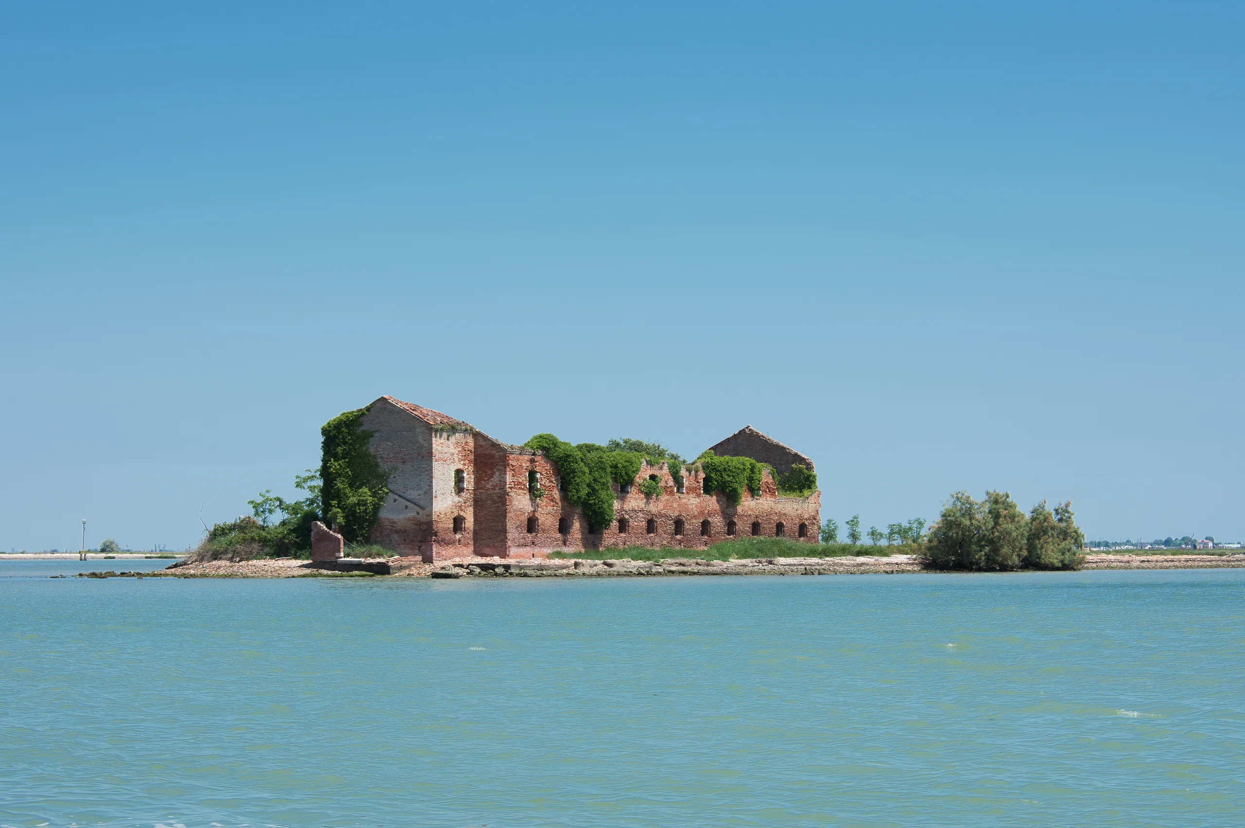 The ruined brick buildings of an abandoned lagoon island standing among trees, seen across flat pale green water.