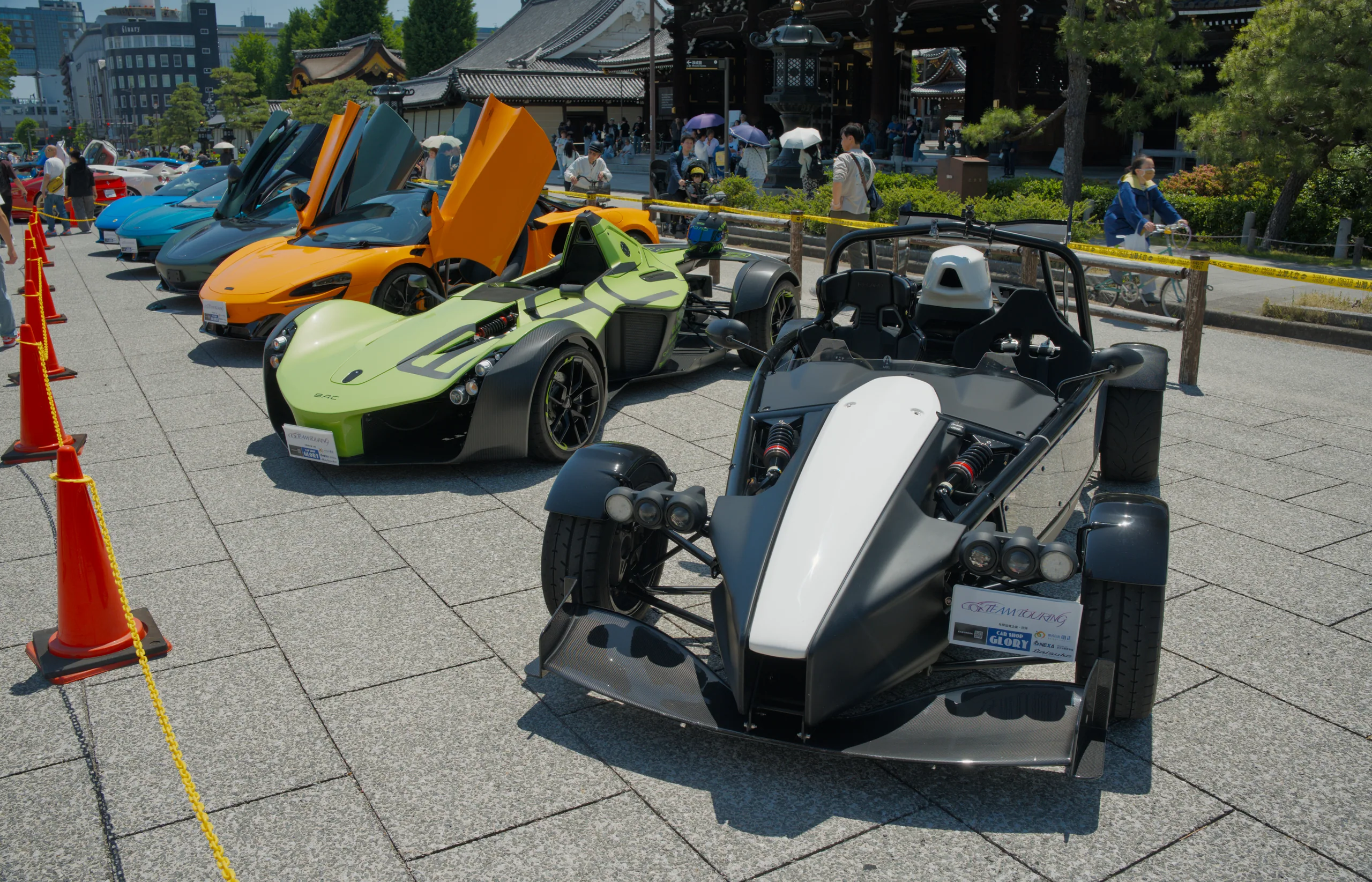 Multiple sports cars of various colors at a paved outdoor car show, including orange, yellow, and light-colored vehicles.