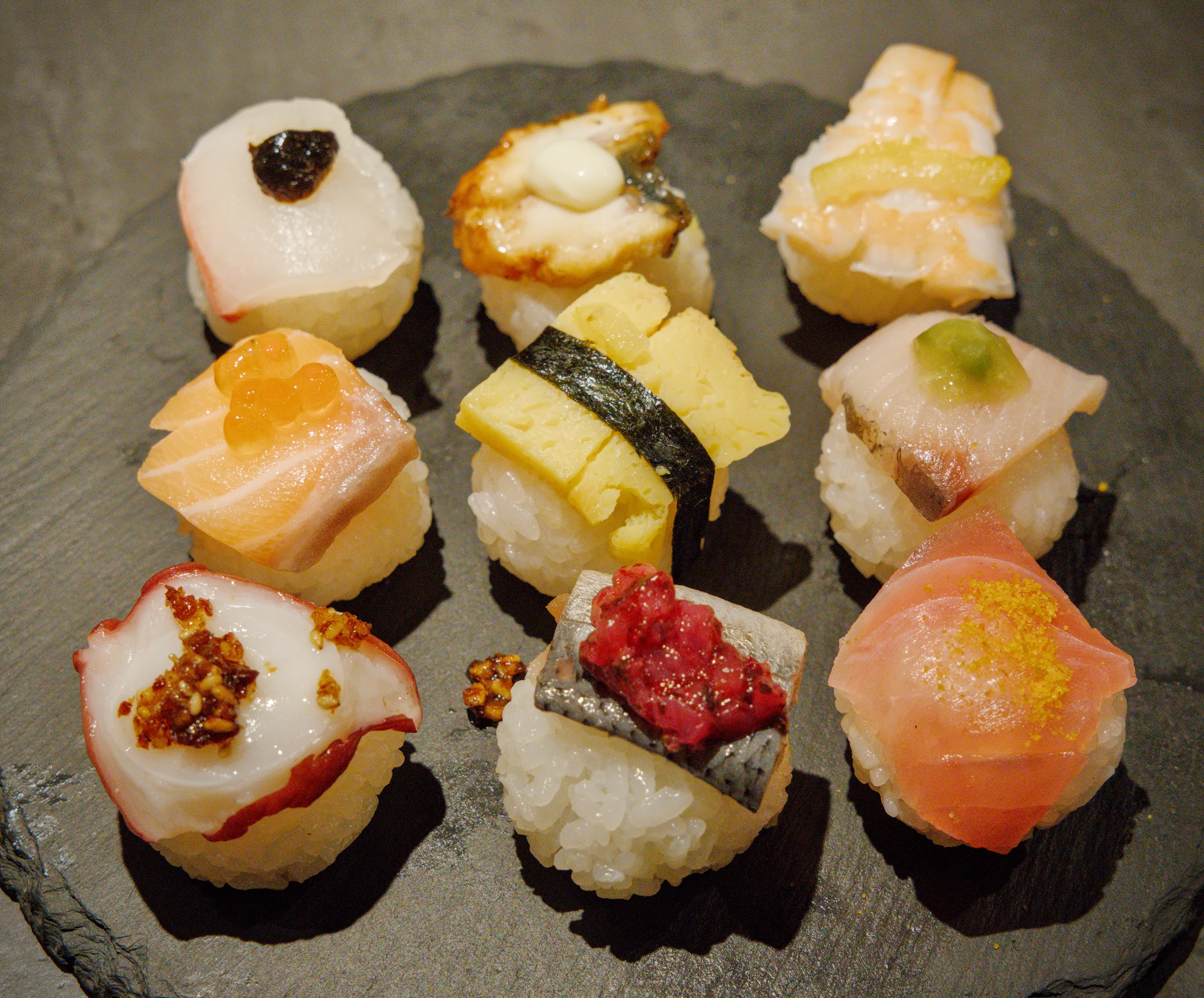 Nine pieces of nigiri sushi with varied toppings arranged on a dark grey plate.