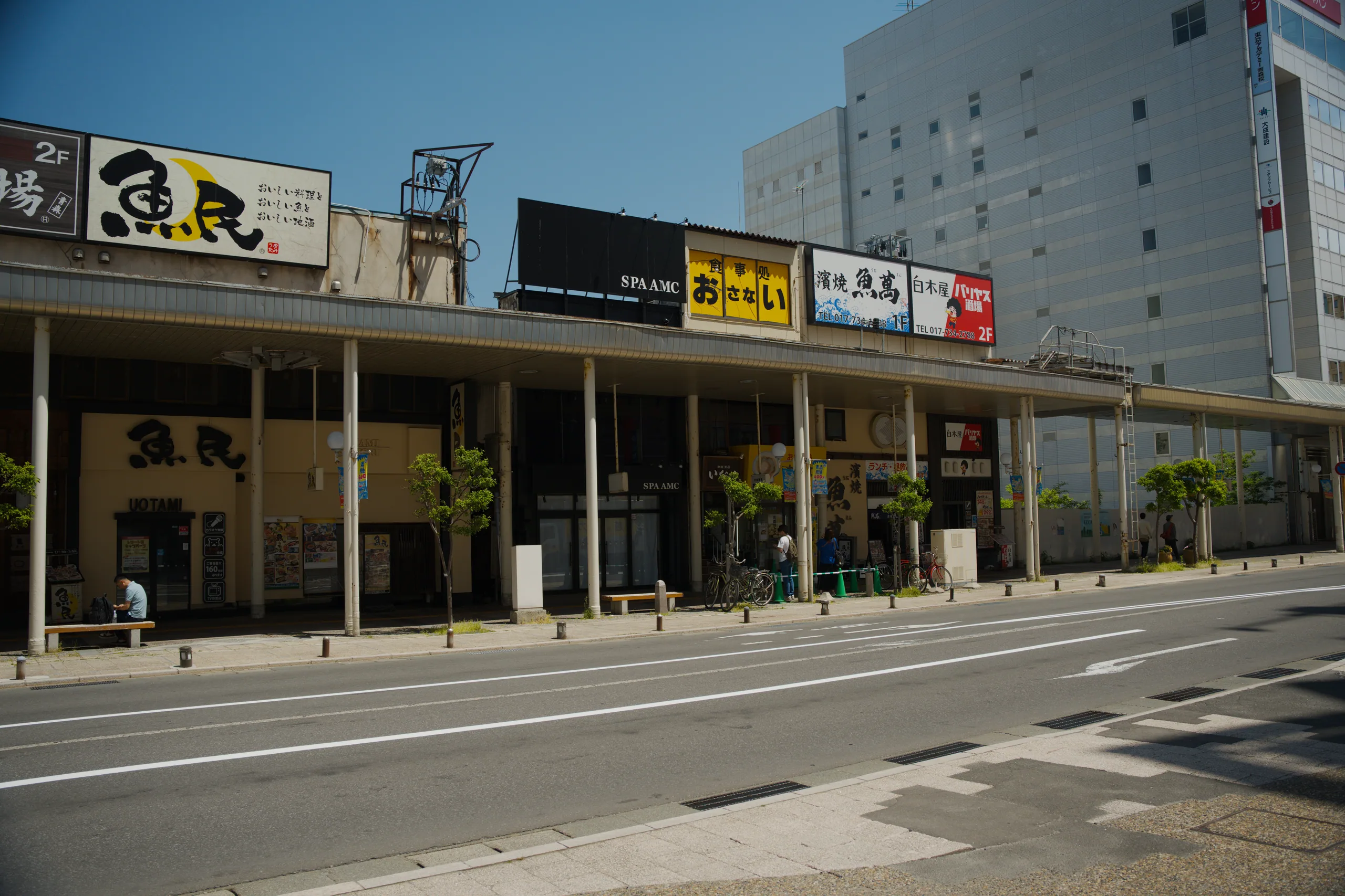 Storefronts with signs line a street in daylight.