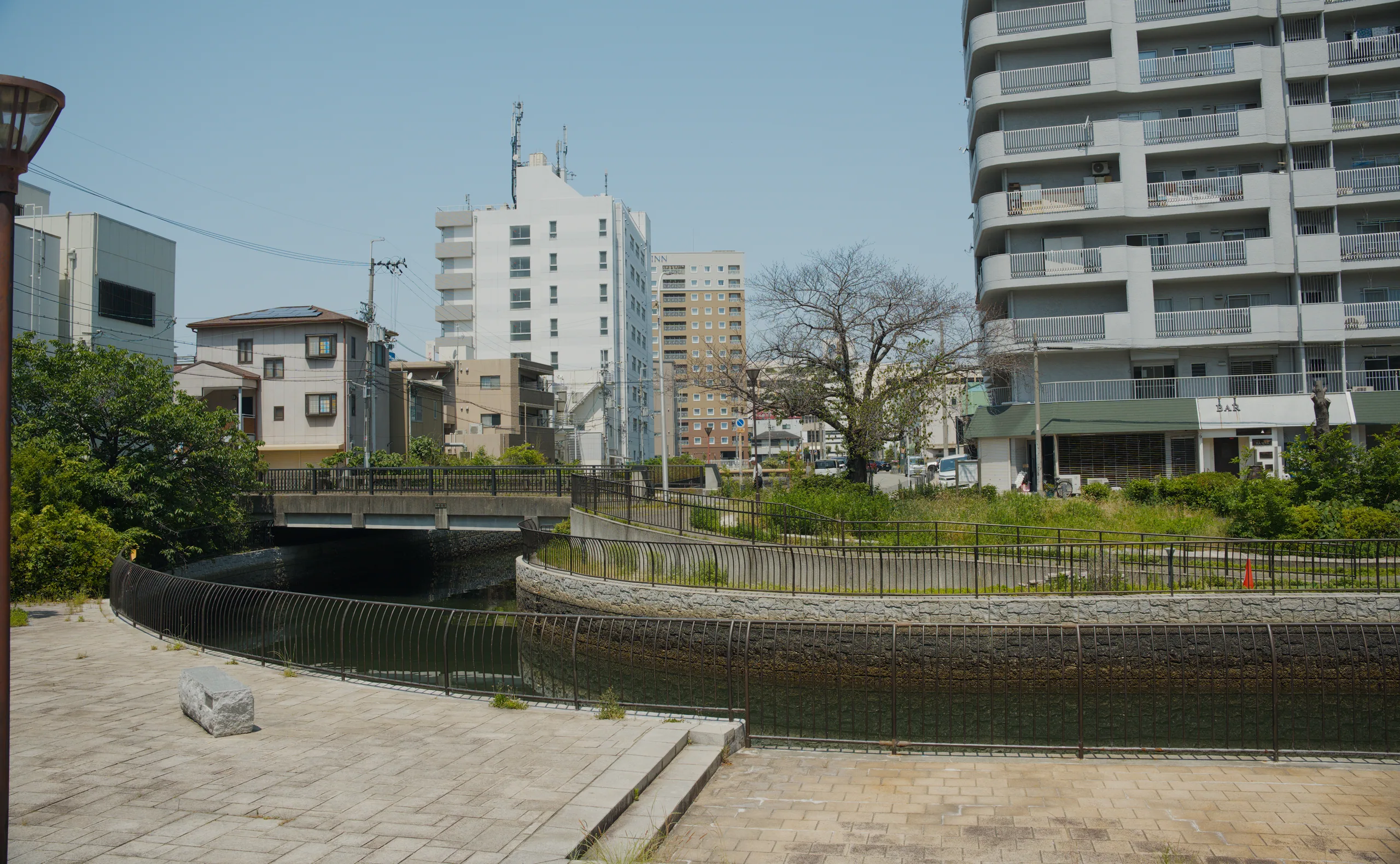 Modern apartment buildings frame a quiet street corner with a small tree growing at the sidewalk's edge.