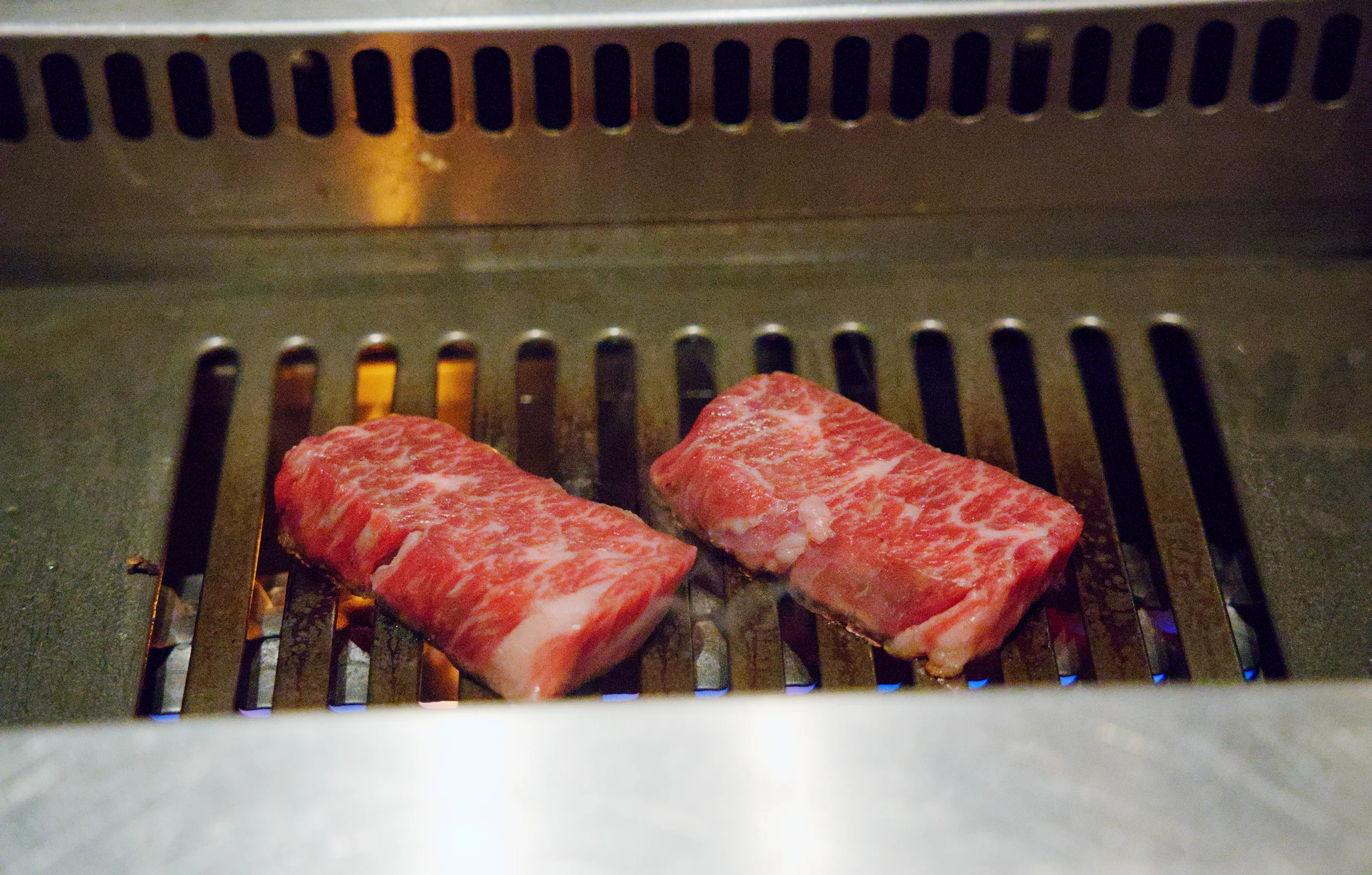 Red meat pieces cook on a metal tabletop grill surrounded by cooking utensils and other table settings.
