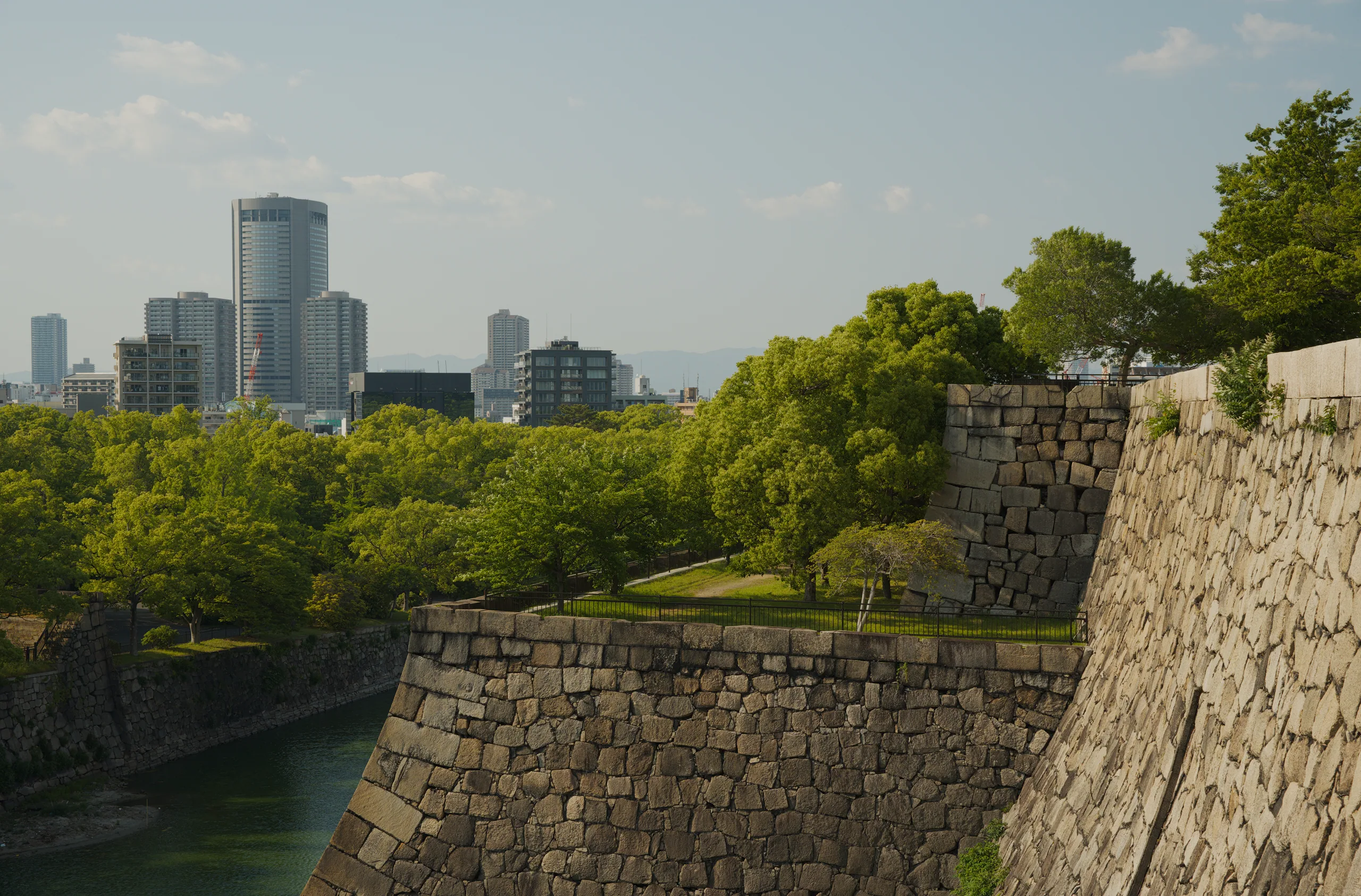 Stone castle fortification walls beside a river with forested banks and a distant city skyline.