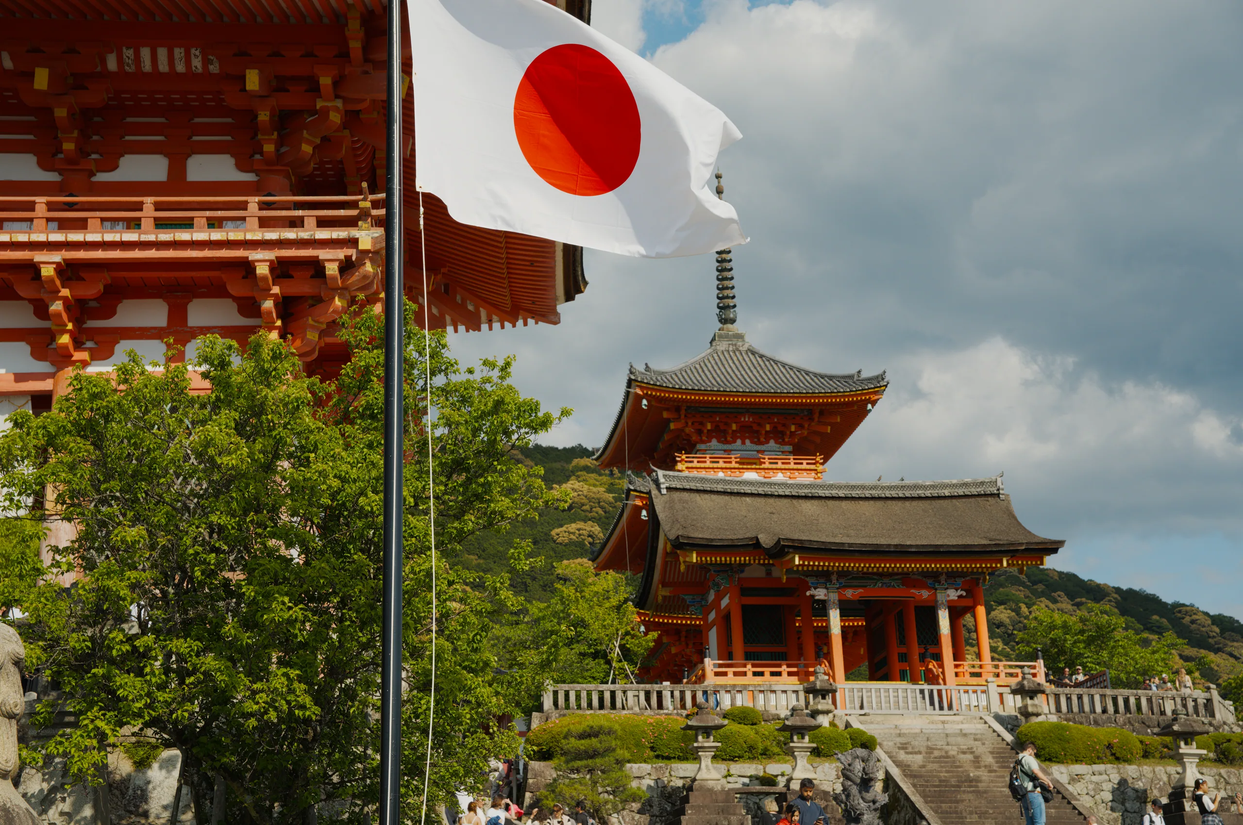 A Japanese flag flies before a tiered red pagoda set on a forested hillside.