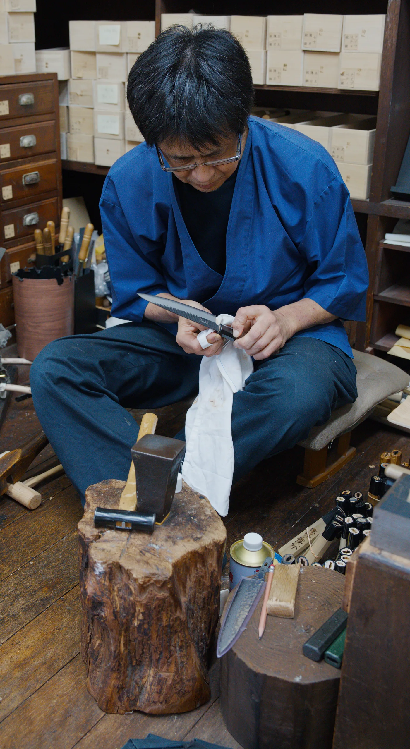 A person in a blue shirt carves a piece of wood while squatting in a workshop.
