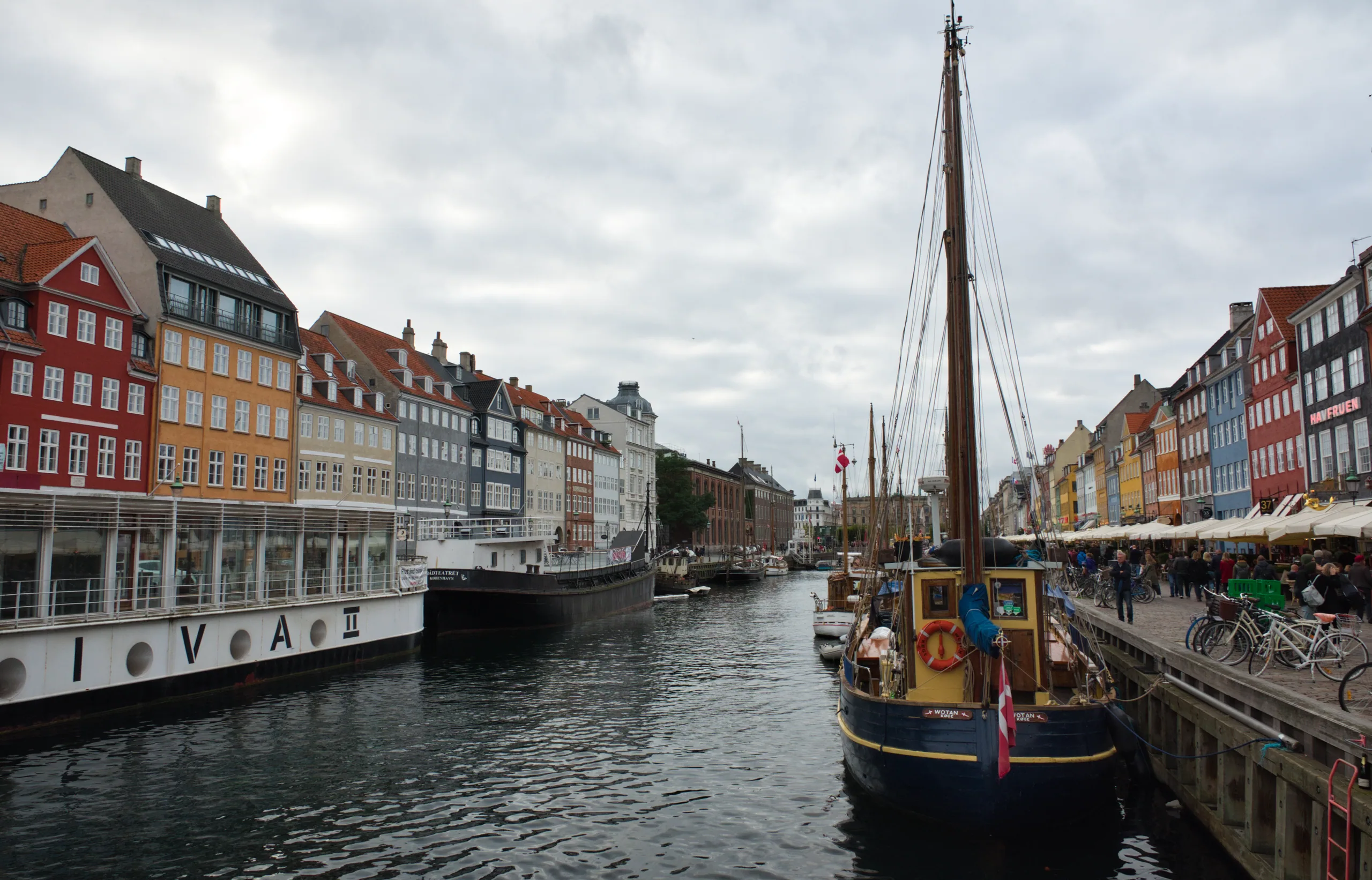 The Nyhavn canal under a grey sky, wooden sailing boats moored along the quay in front of a terrace of narrow red, yellow and ochre townhouses.