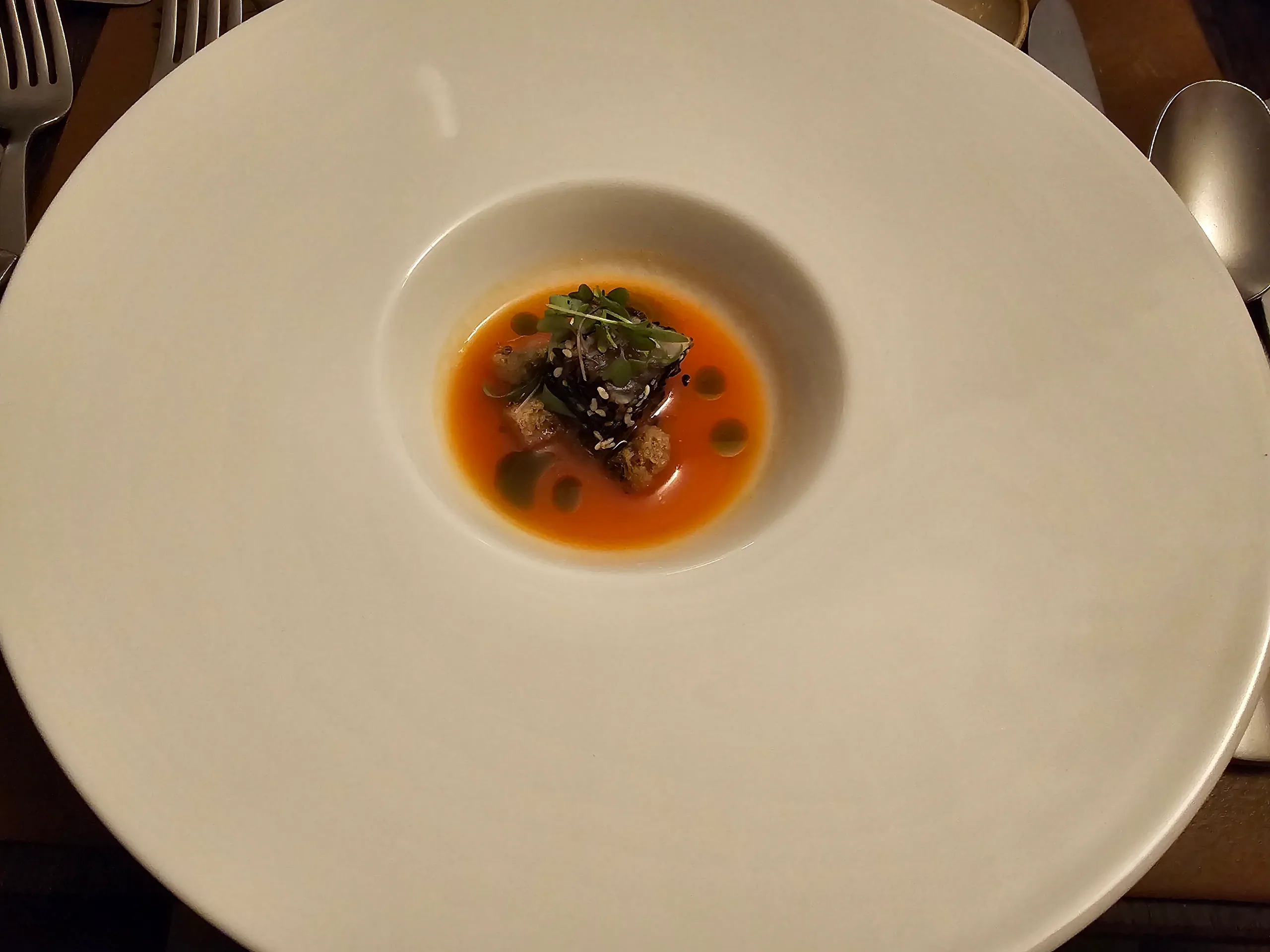 A small restaurant course in the centre of a wide white plate, a fritter in orange broth with herbs and green oil.
