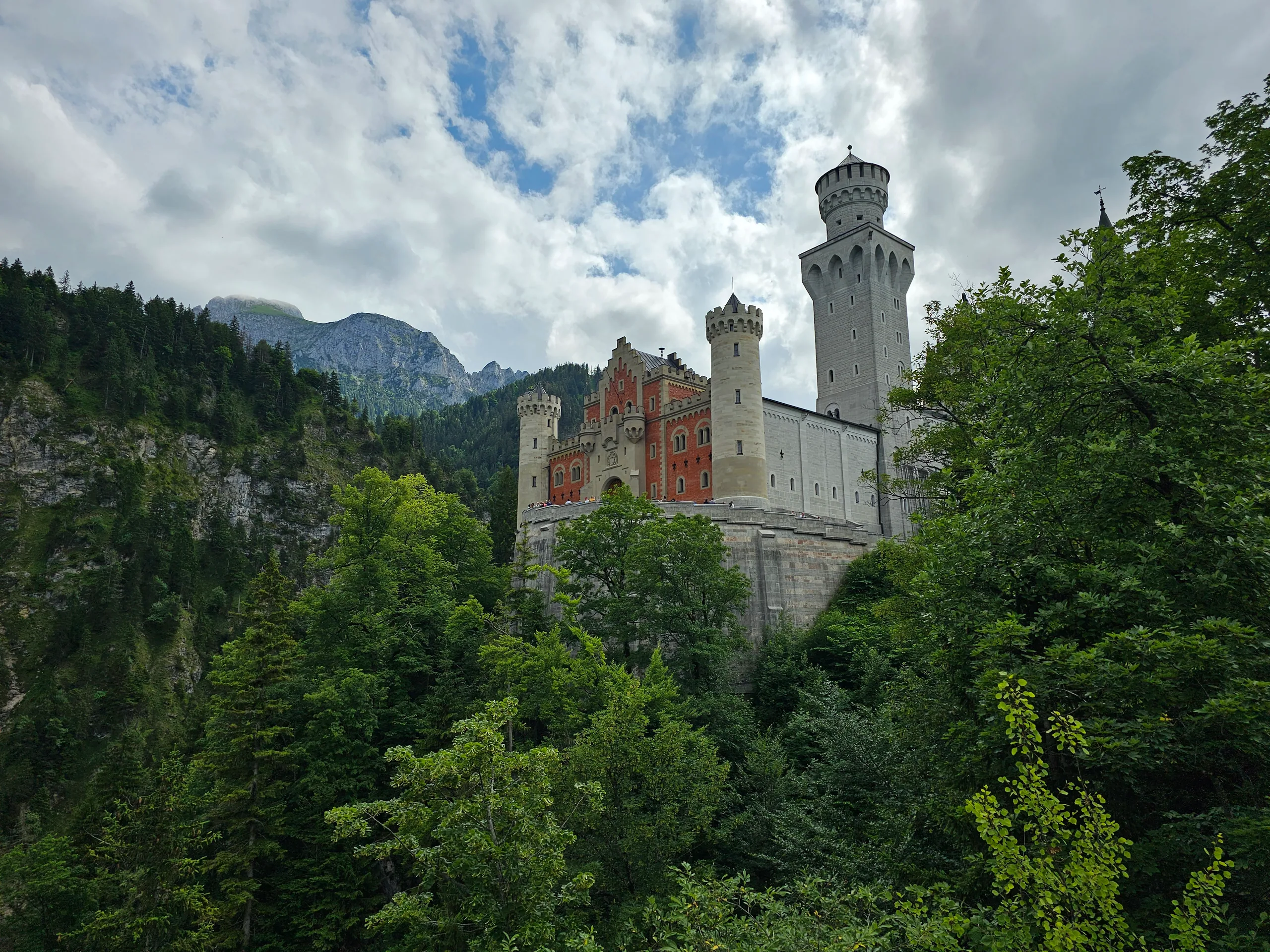 Neuschwanstein castle on its wooded crag, white walls and towers against forested slopes and cloud.