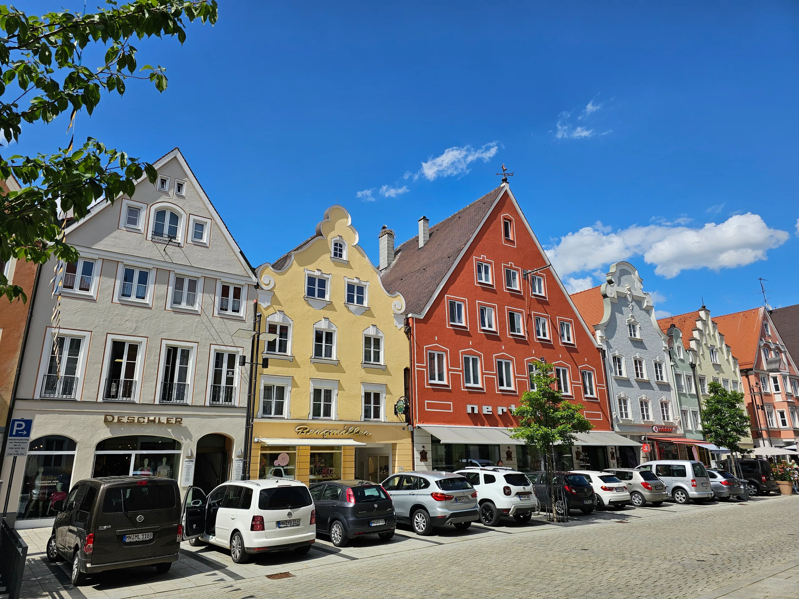 A row of stepped-gable townhouses in grey, yellow and red along a sunlit street, cars parked in front.