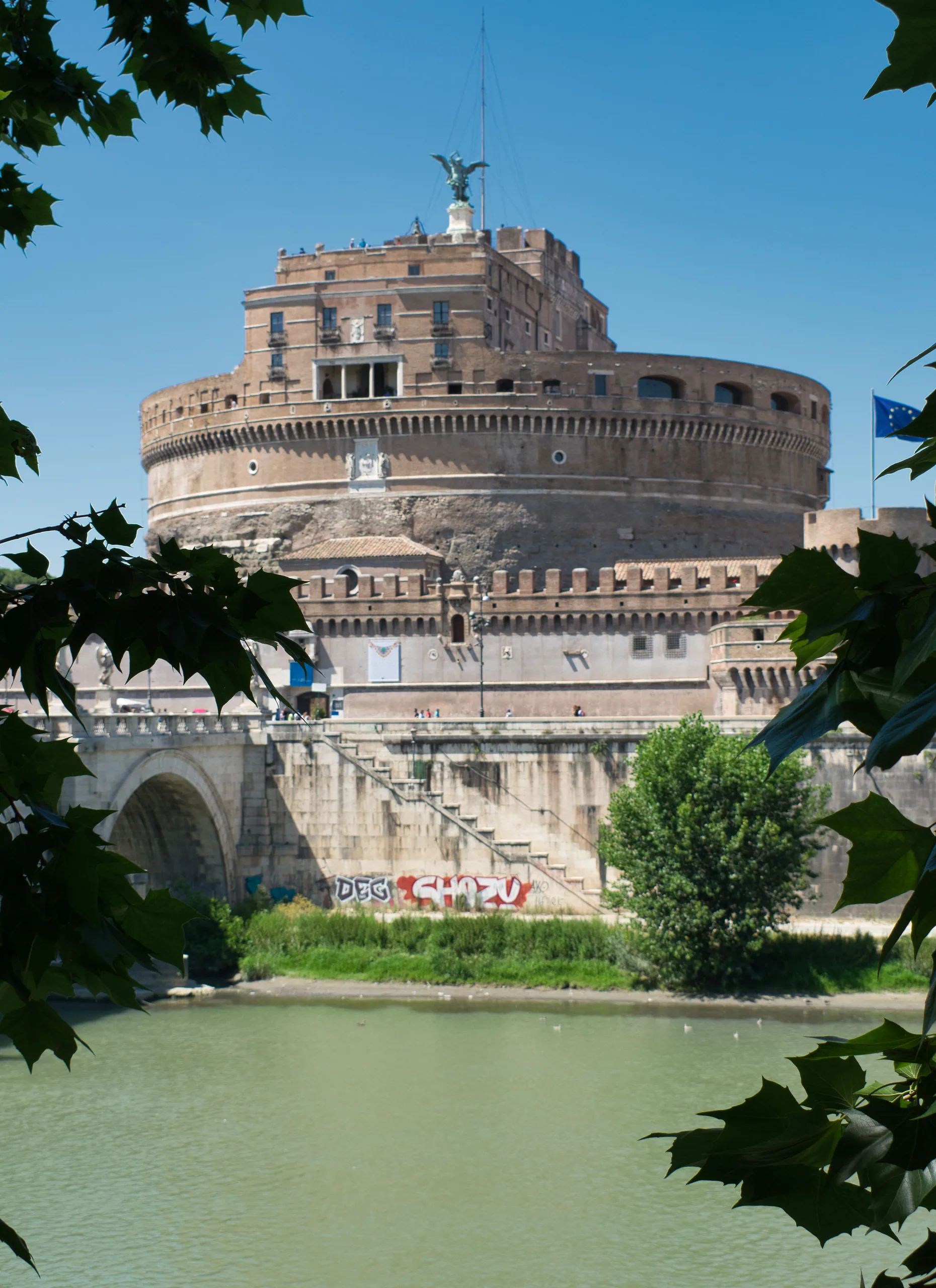 Castel Sant'Angelo across the Tiber, framed by plane tree leaves, with graffiti along the embankment wall below.