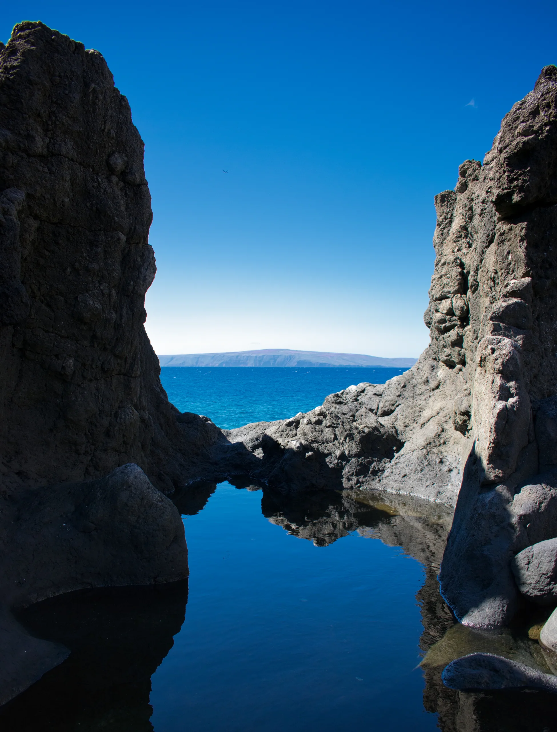 A dark tidal pool between two lava headlands, reflecting the sky, with the ocean and a distant island beyond.