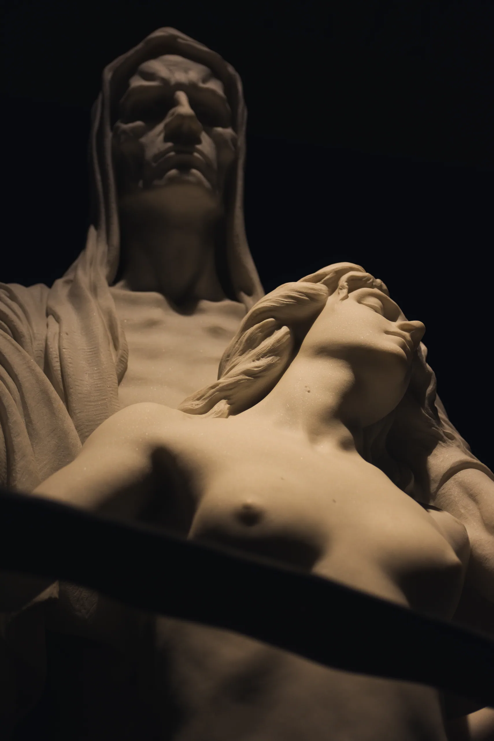 A pale marble sculpture of a veiled figure standing over a reclining nude, lit dramatically against a black background.
