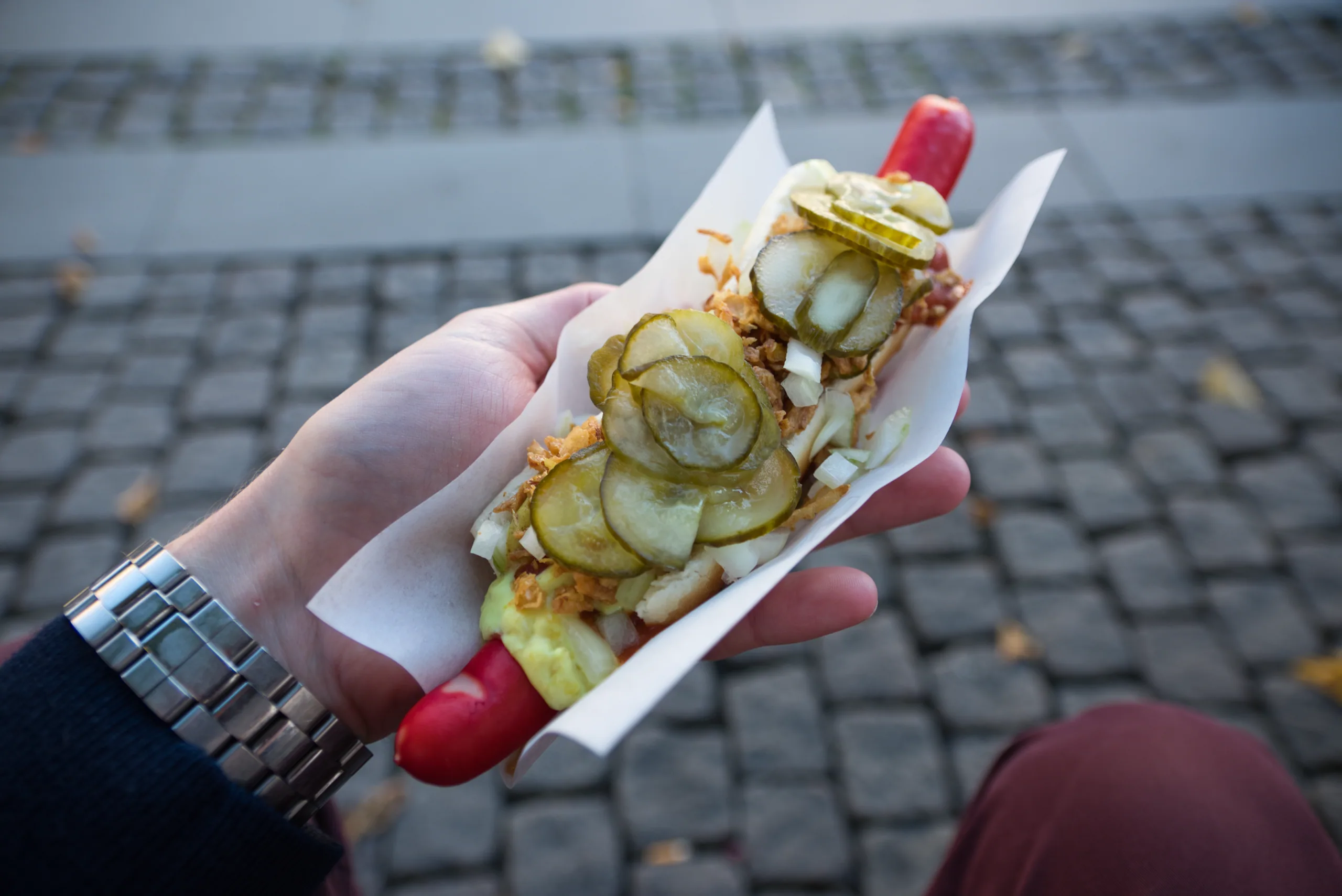 A hot dog held in one hand over cobblestones, loaded with pickled cucumber, crisp fried onion and remoulade.