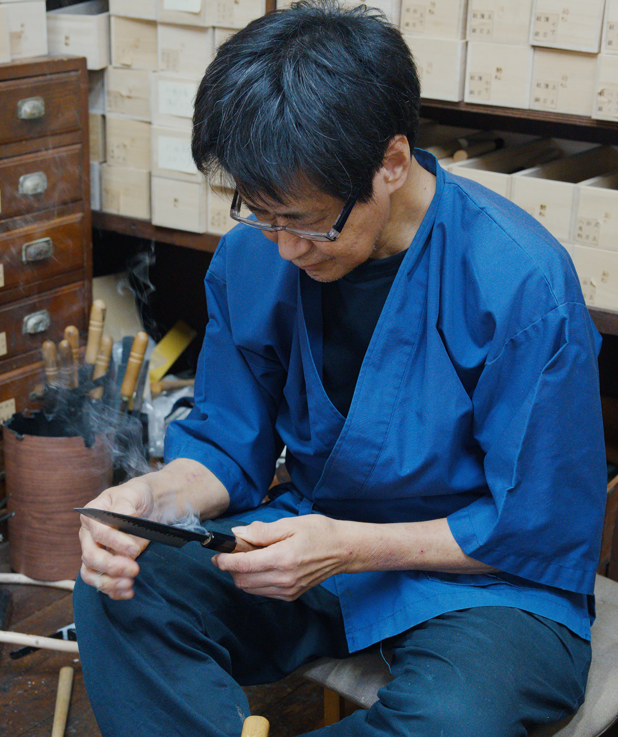 A person in a blue shirt works with wood shavings at a workshop desk with storage drawers visible behind them.