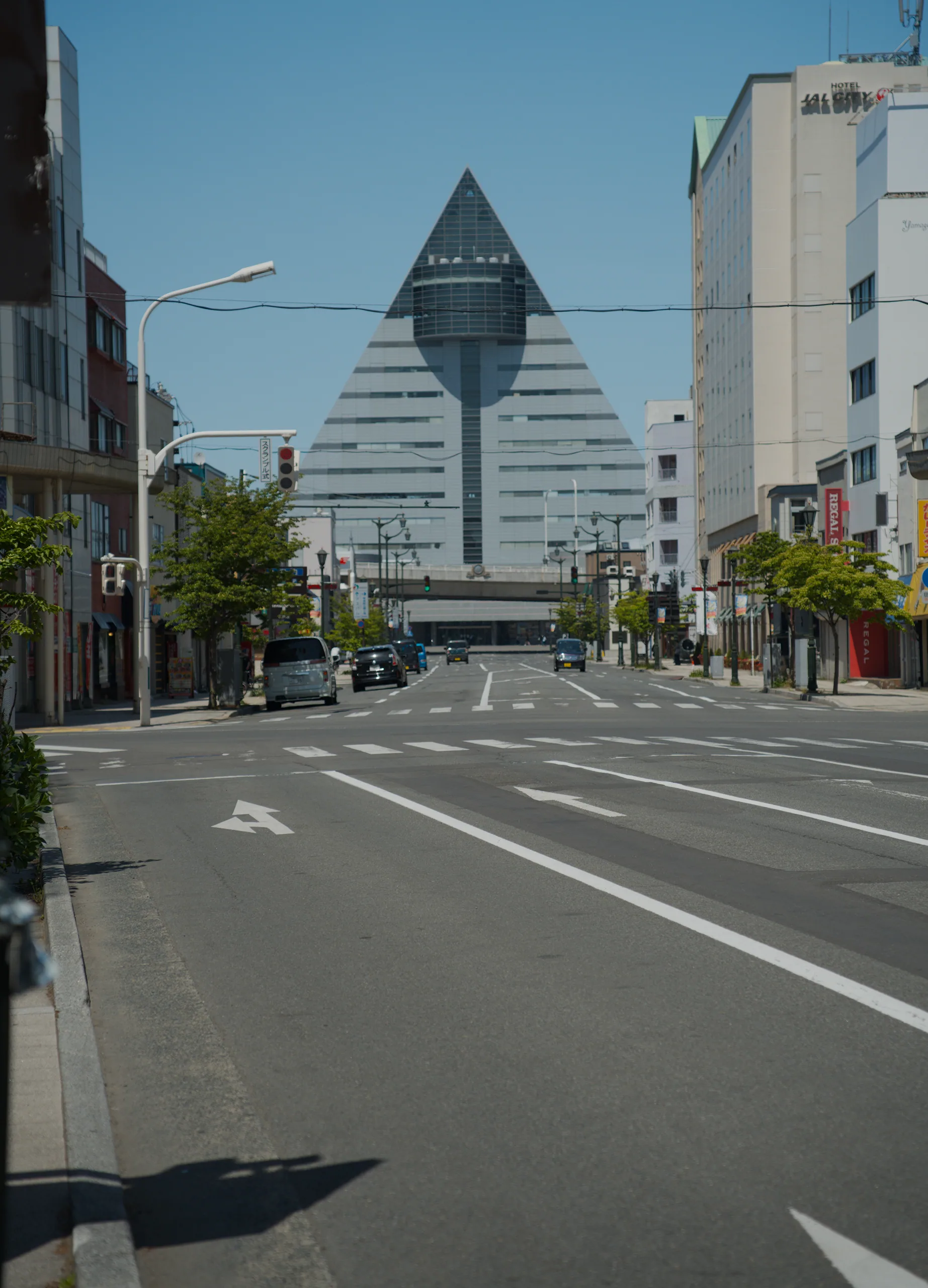 A striped pyramid-shaped building rises above a Japanese street lined with smaller buildings and trees.