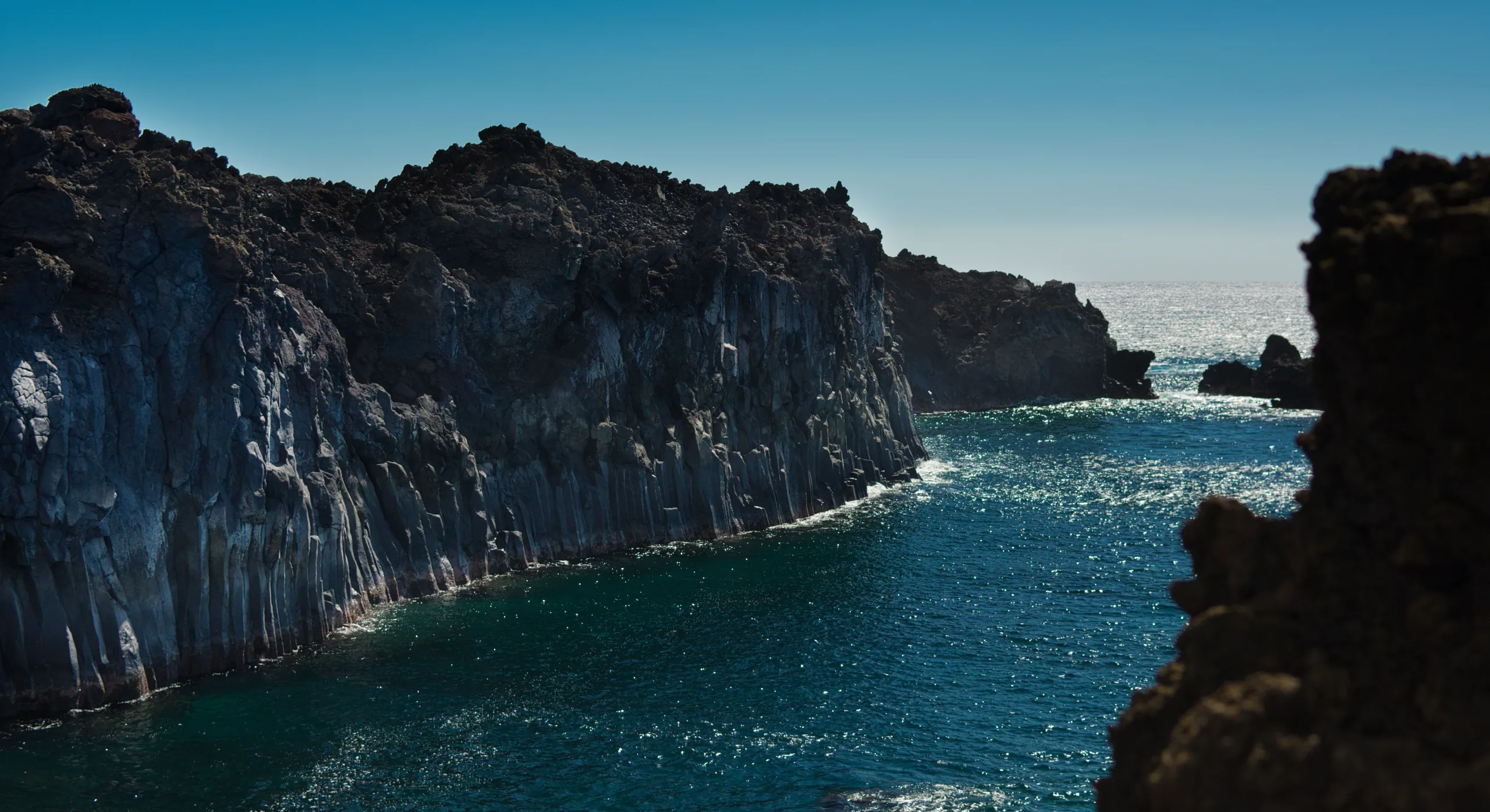 Columnar sea cliffs of dark basalt dropping straight into clear blue water along a Hawaiian coast.
