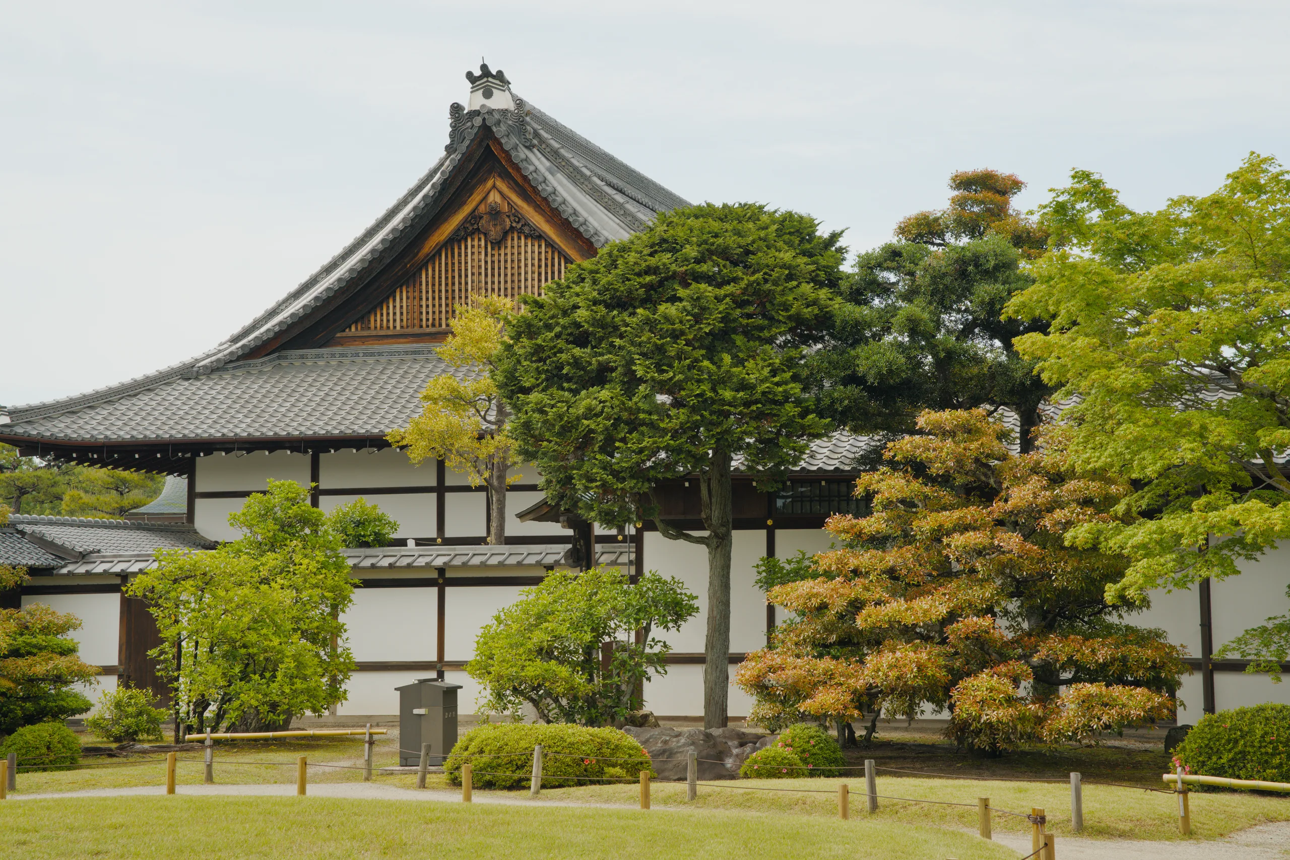 A traditional Japanese building with an upturned roof sits beside a moat lined with stone castle walls, flanked by manicured trees.
