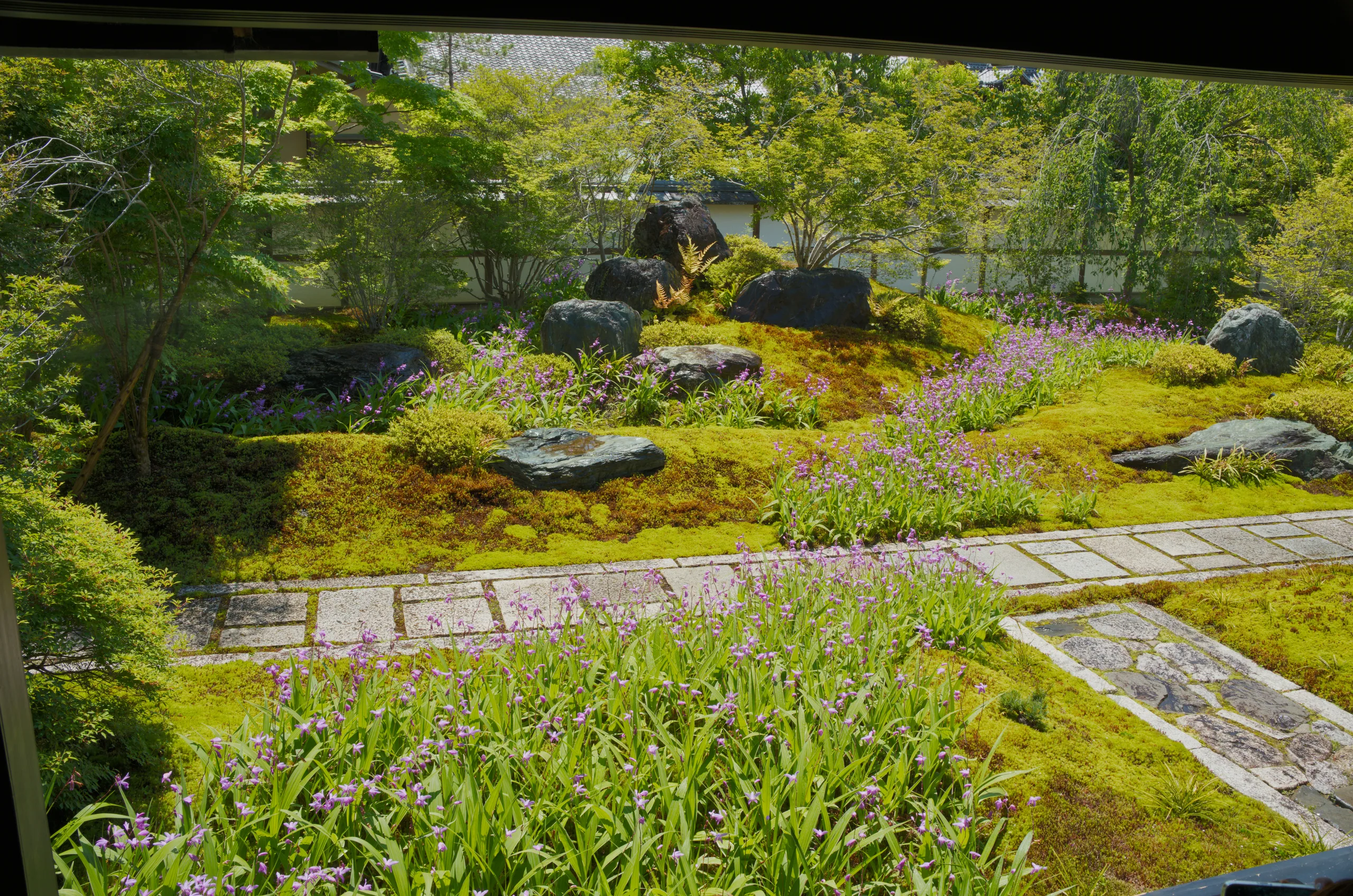 A stepping-stone pathway winds through a moss and grass garden with a stream running alongside lush plantings.