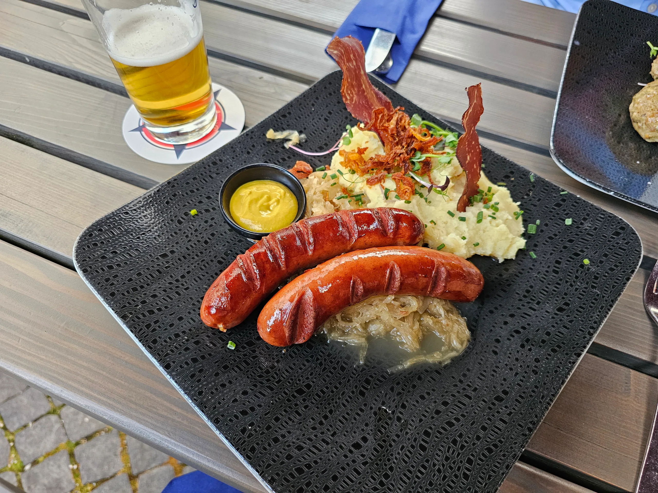 Two grilled sausages with potato salad, crisp bacon and mustard on a slate, a glass of beer behind.