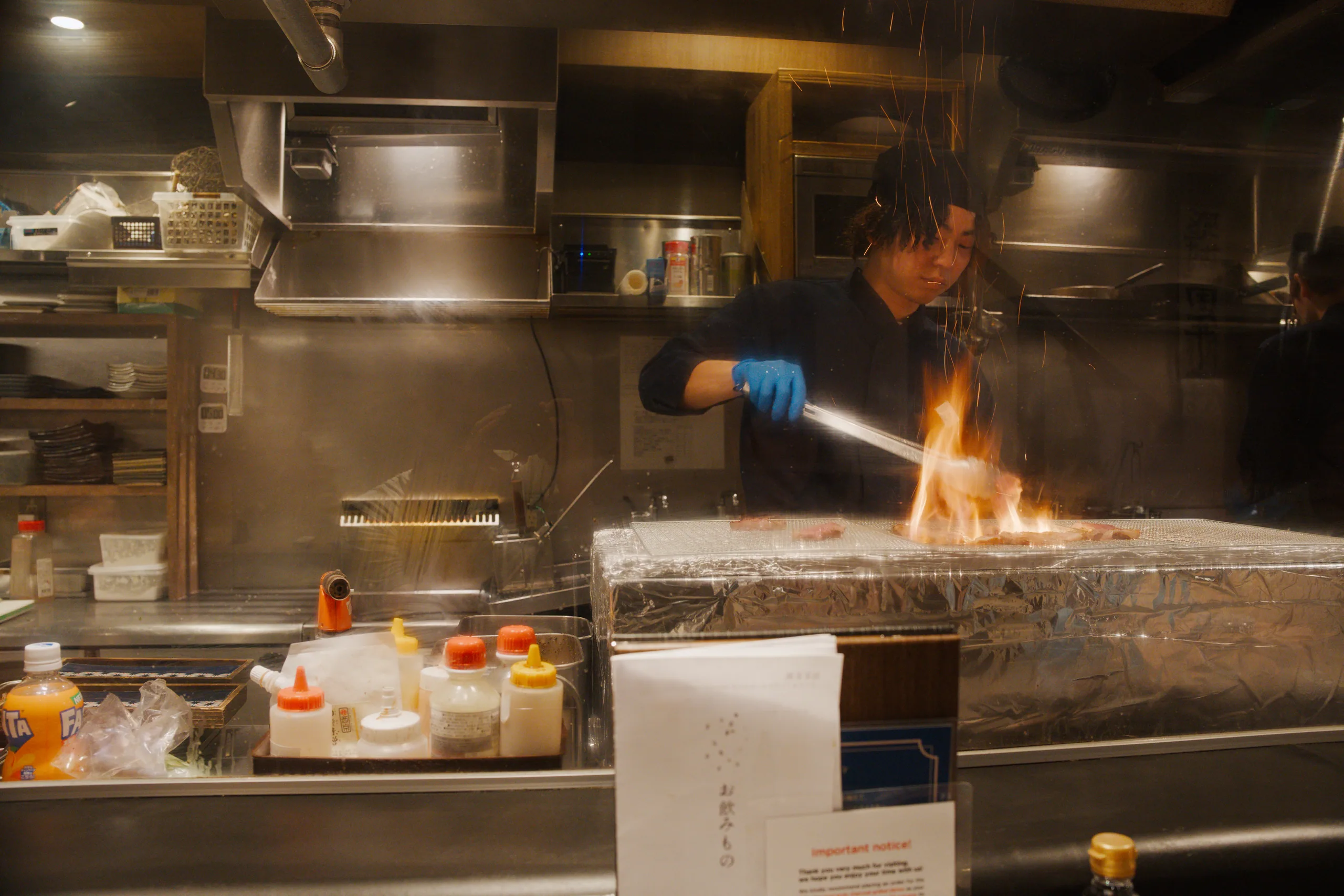 A chef in dark clothes and blue gloves works with flame at a stainless steel counter in an open kitchen.
