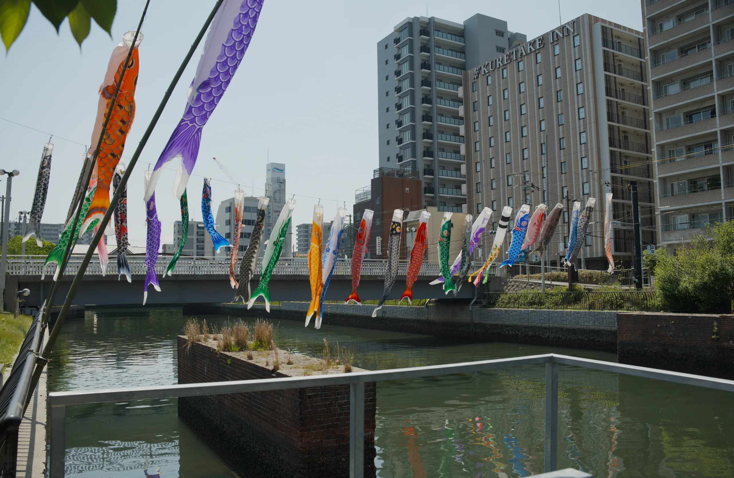 Colorful decorative flags displayed along a waterfront plaza overlooking a canal with modern buildings beyond.