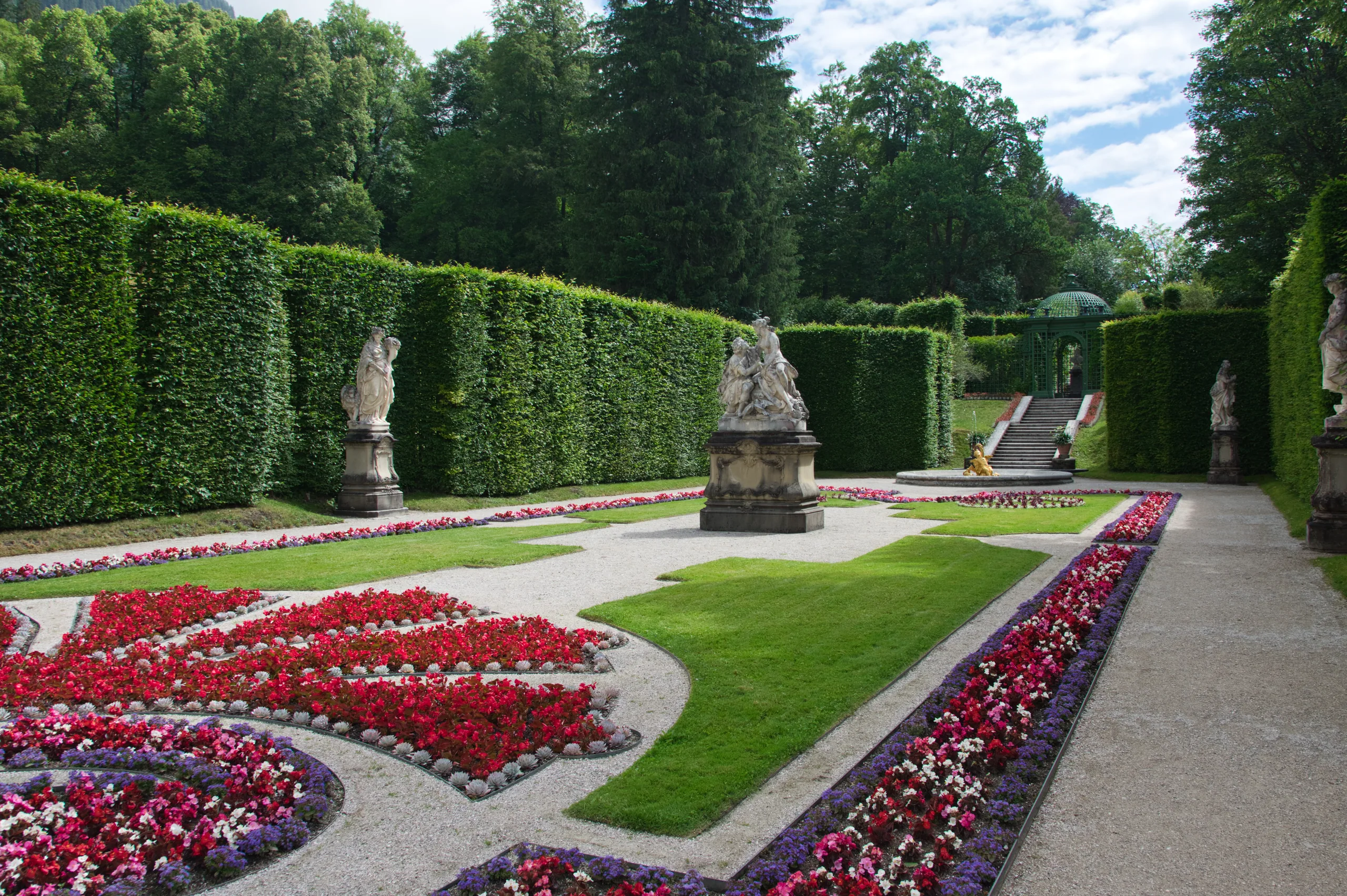 The formal parterre at Linderhof, beds of red and purple flowers around clipped lawn, statues on plinths against tall hedges.