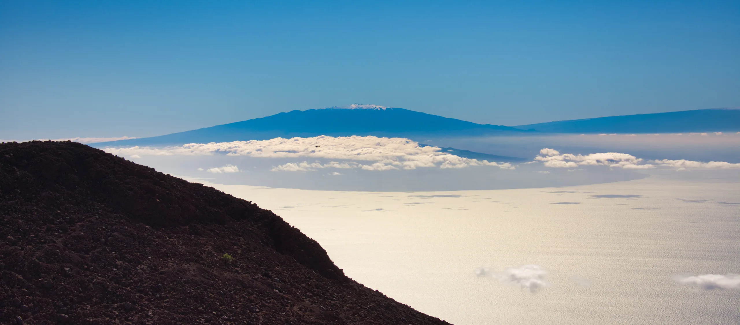 A dark cinder slope falling away to a flat white sea of cloud, a distant volcano rising above it.