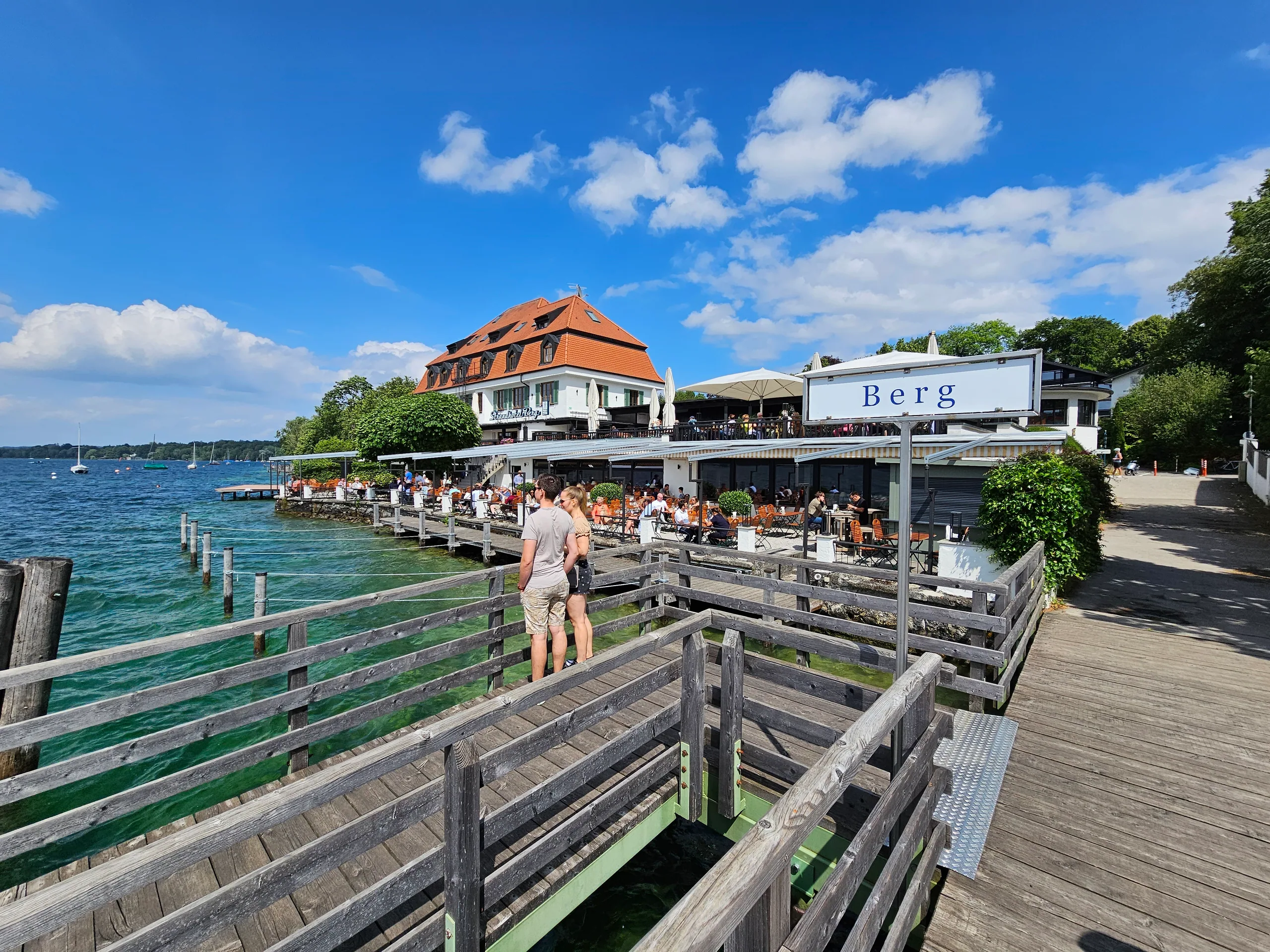 A lakeside beer garden at Berg on the Starnberger See, wooden jetties running out into clear green water under a bright sky.