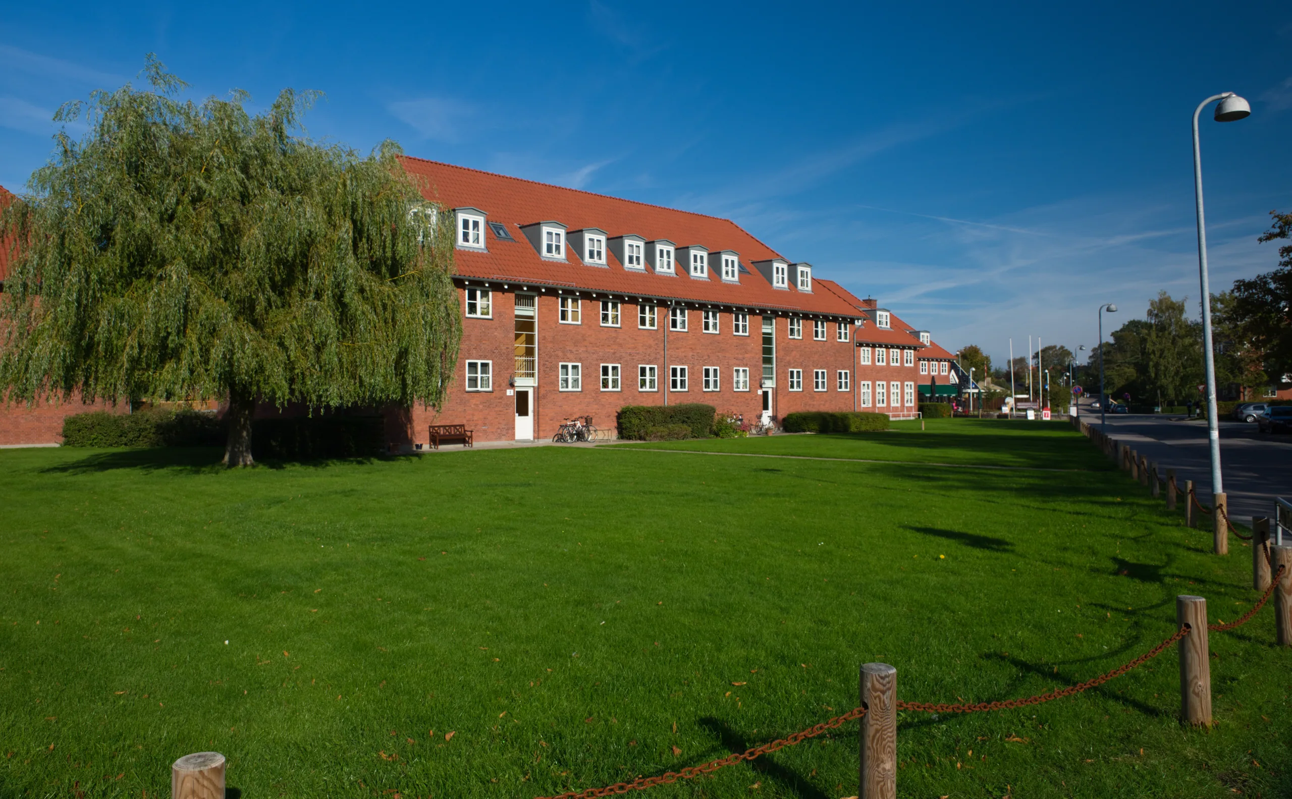 A long red-brick building with a red tiled roof behind a mown lawn and a weeping willow, under a clear blue sky.