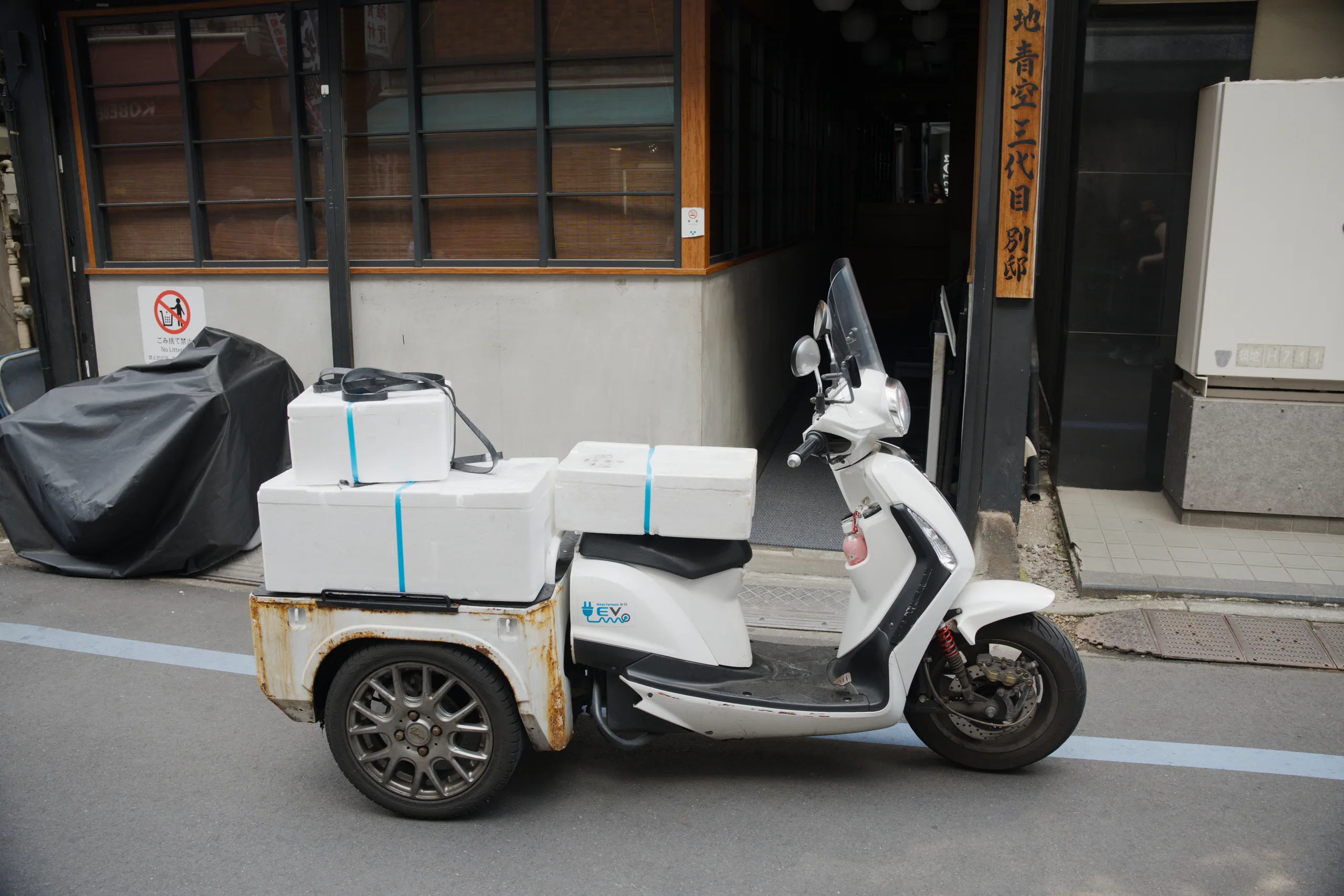 A white cargo scooter with delivery box parked on a narrow urban street.