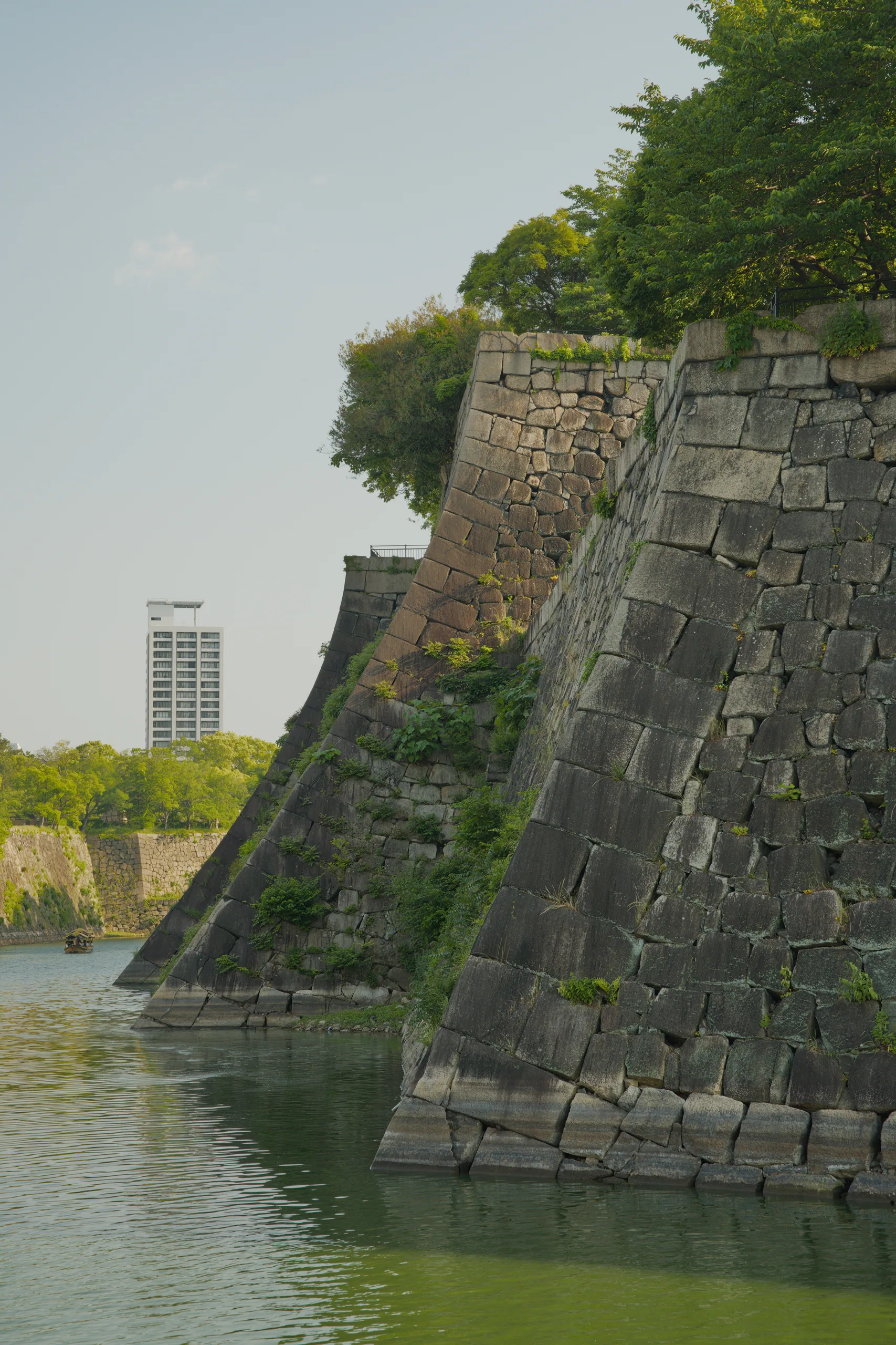 Stone castle fortification walls sloping steeply toward water, with vegetation at the crest and a modern building visible inland.