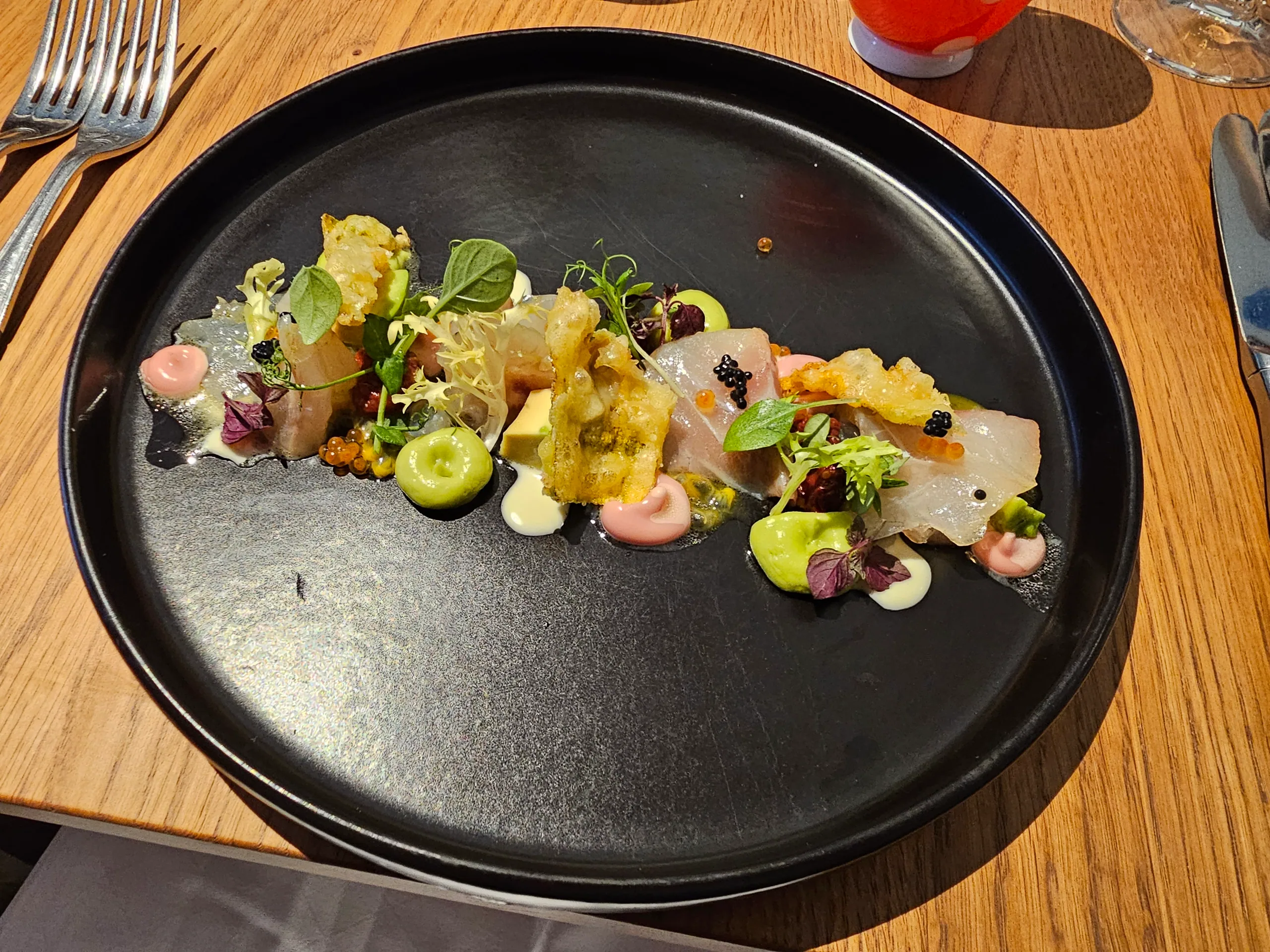 A restaurant course of cured fish with pickled vegetables, herbs and dots of sauce on a black plate.
