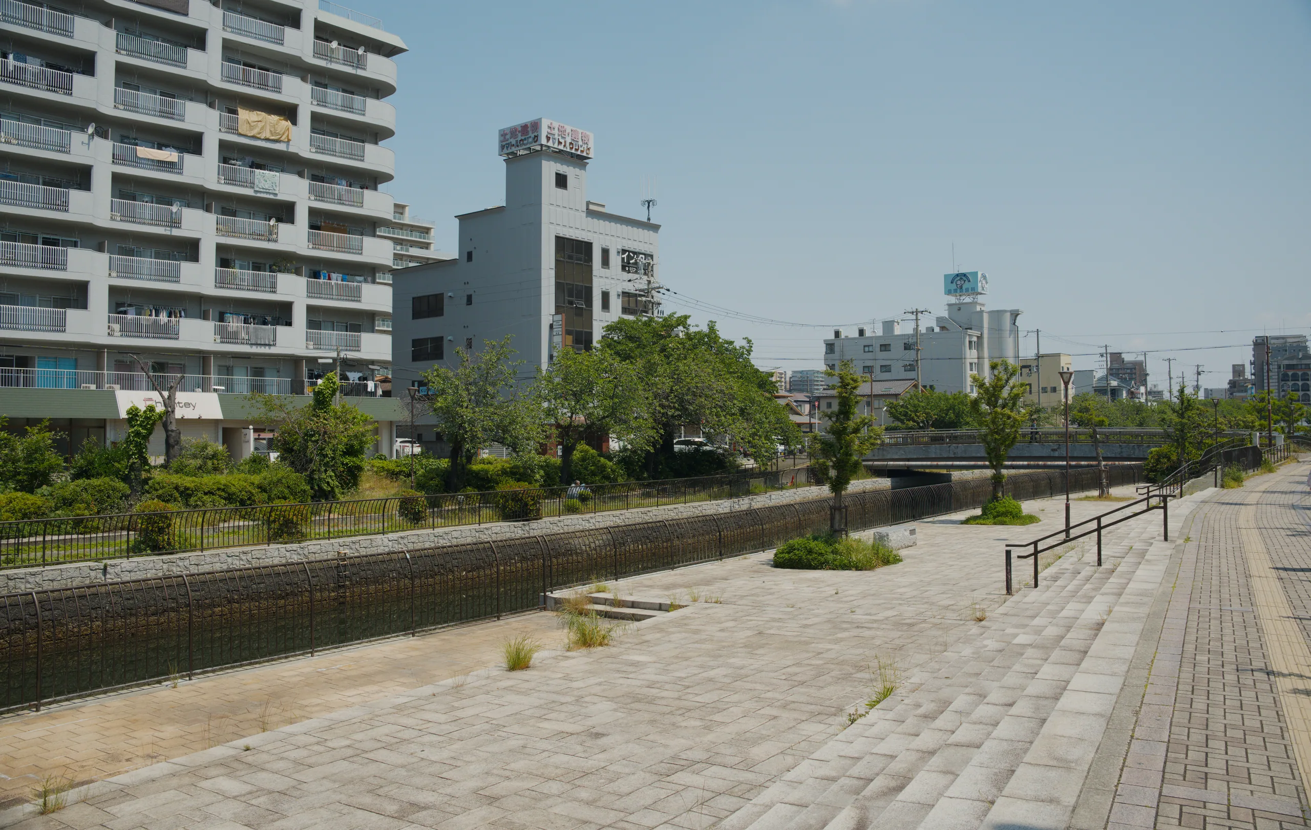 A paved promenade runs alongside an urban river lined with modern apartment buildings and metal railings.