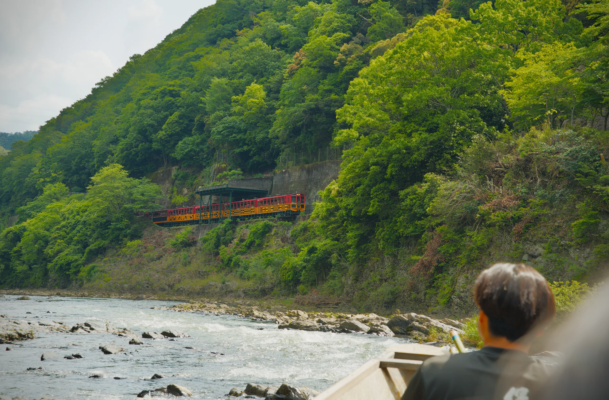 Person seated on a rocky riverbank with steep forested hills and river water across.