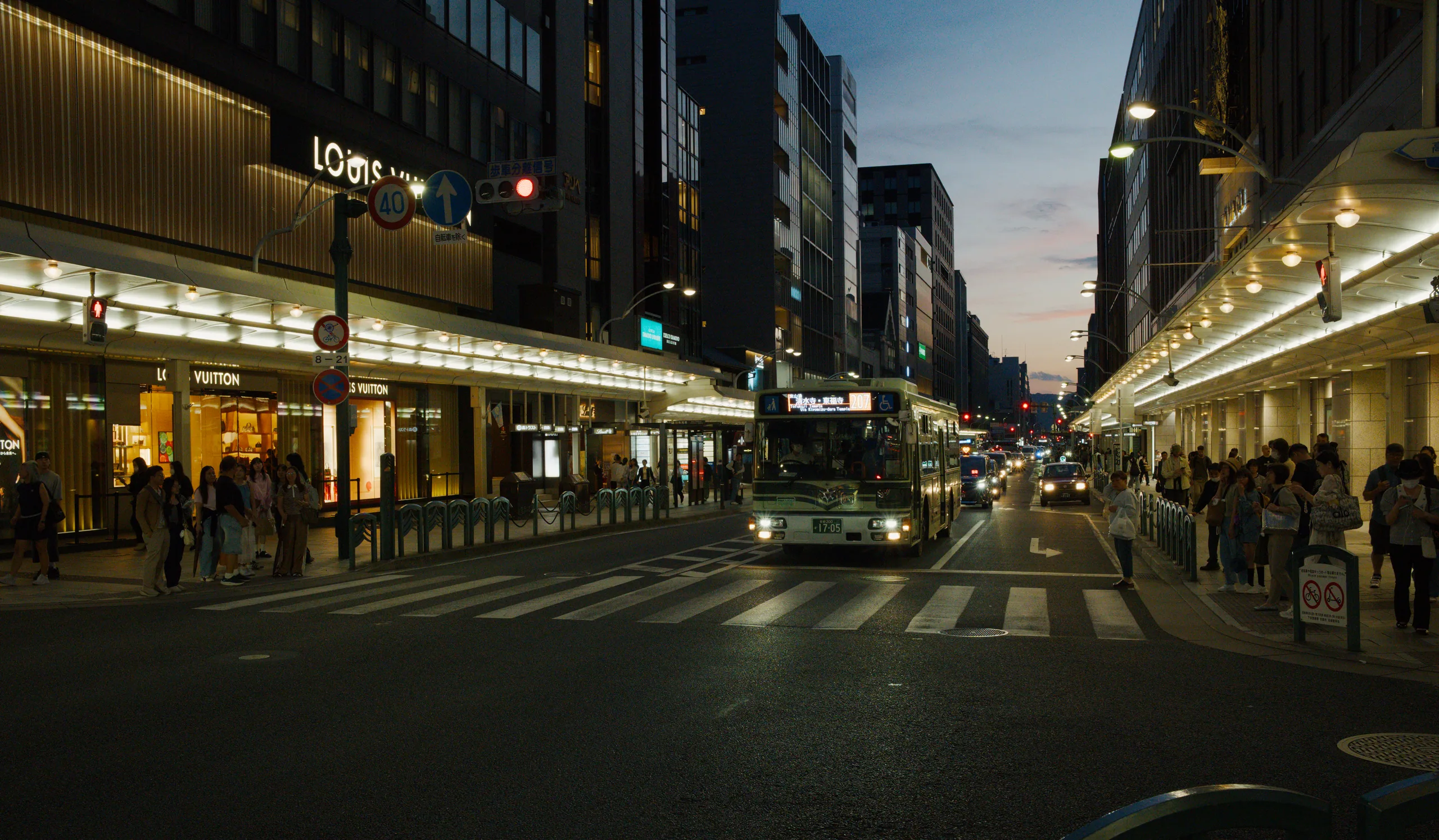 An illuminated city street with tall buildings, lit storefronts, and pedestrians crossing.