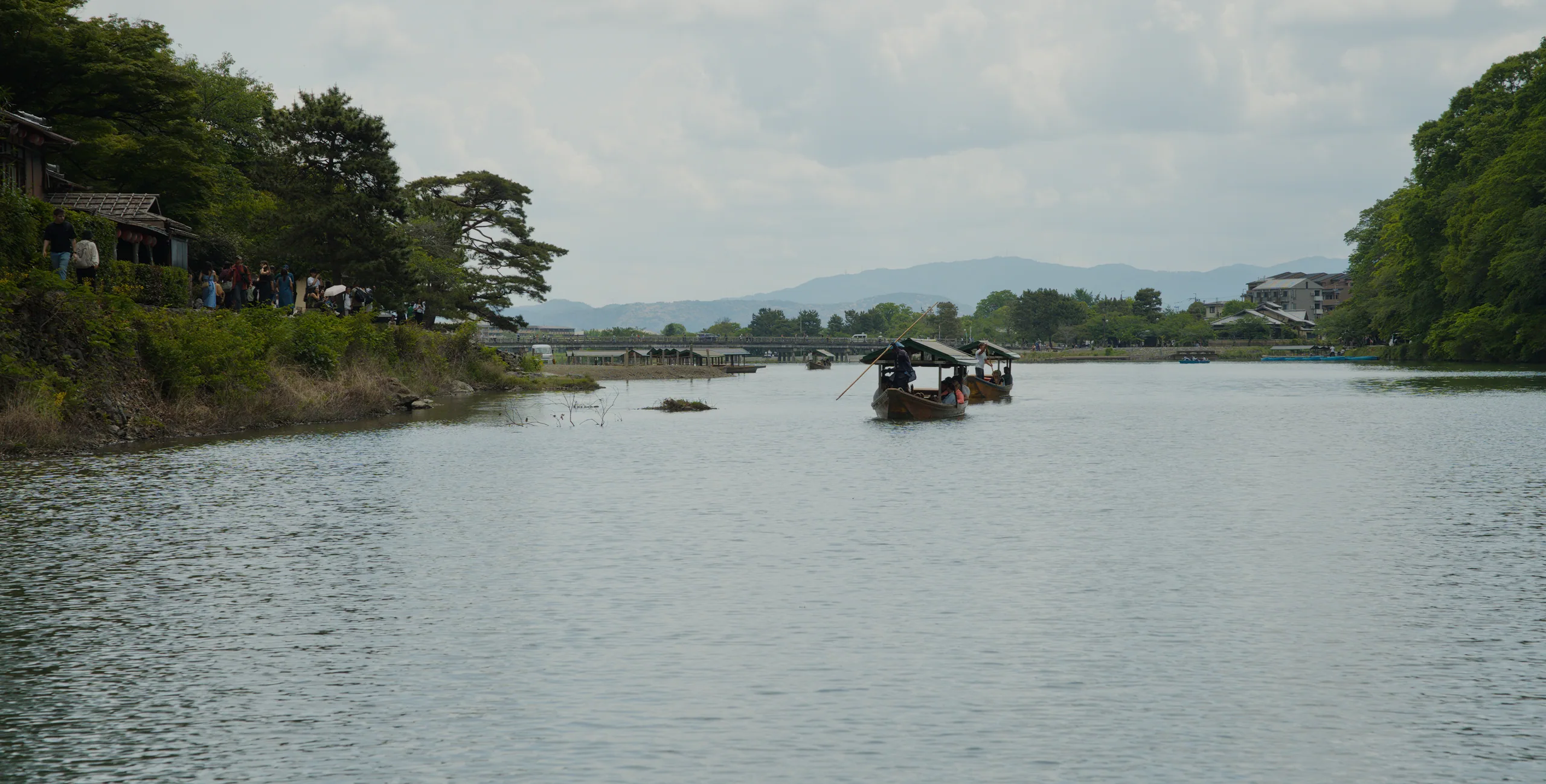 A dark boat on a wide river runs between forested hillsides under overcast sky.