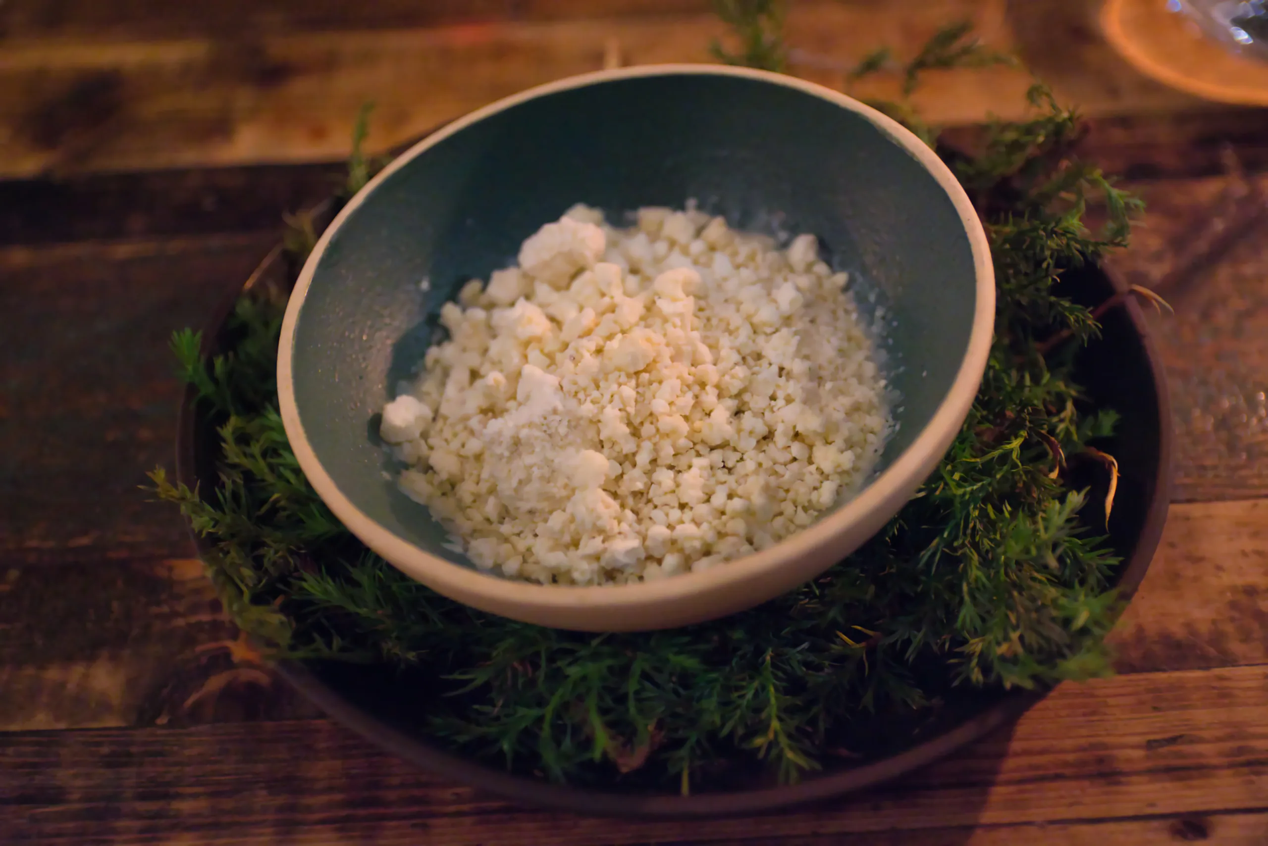 A bowl of crumbled white curd set inside a second dish packed with green herbs, on a wooden table.