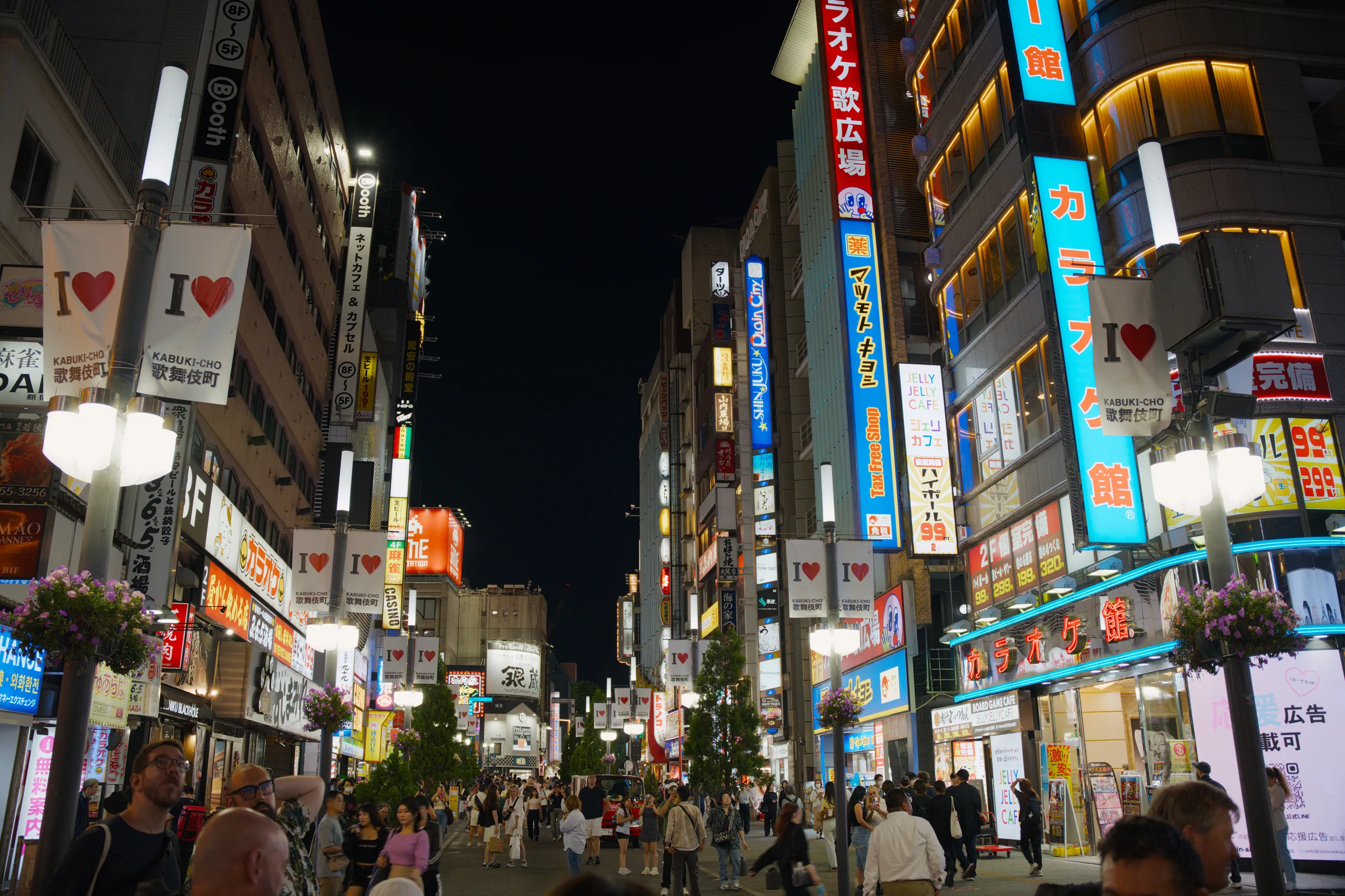Wide pedestrian shopping street at night filled with crowds beneath tall buildings covered in colorful lit signs and digital displays.