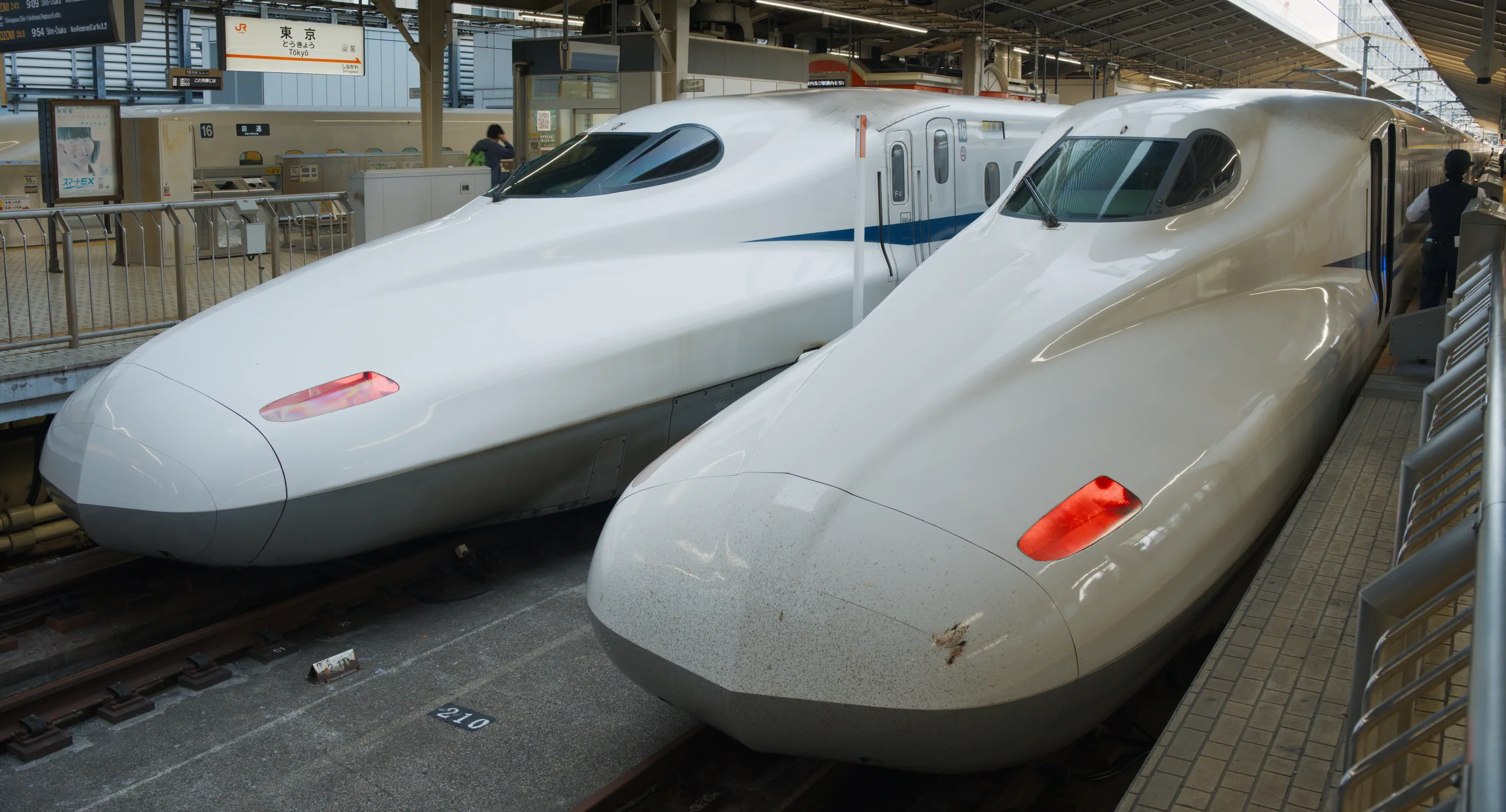 White bullet trains rest at a train station platform with metal barriers and a patterned geometric ceiling.