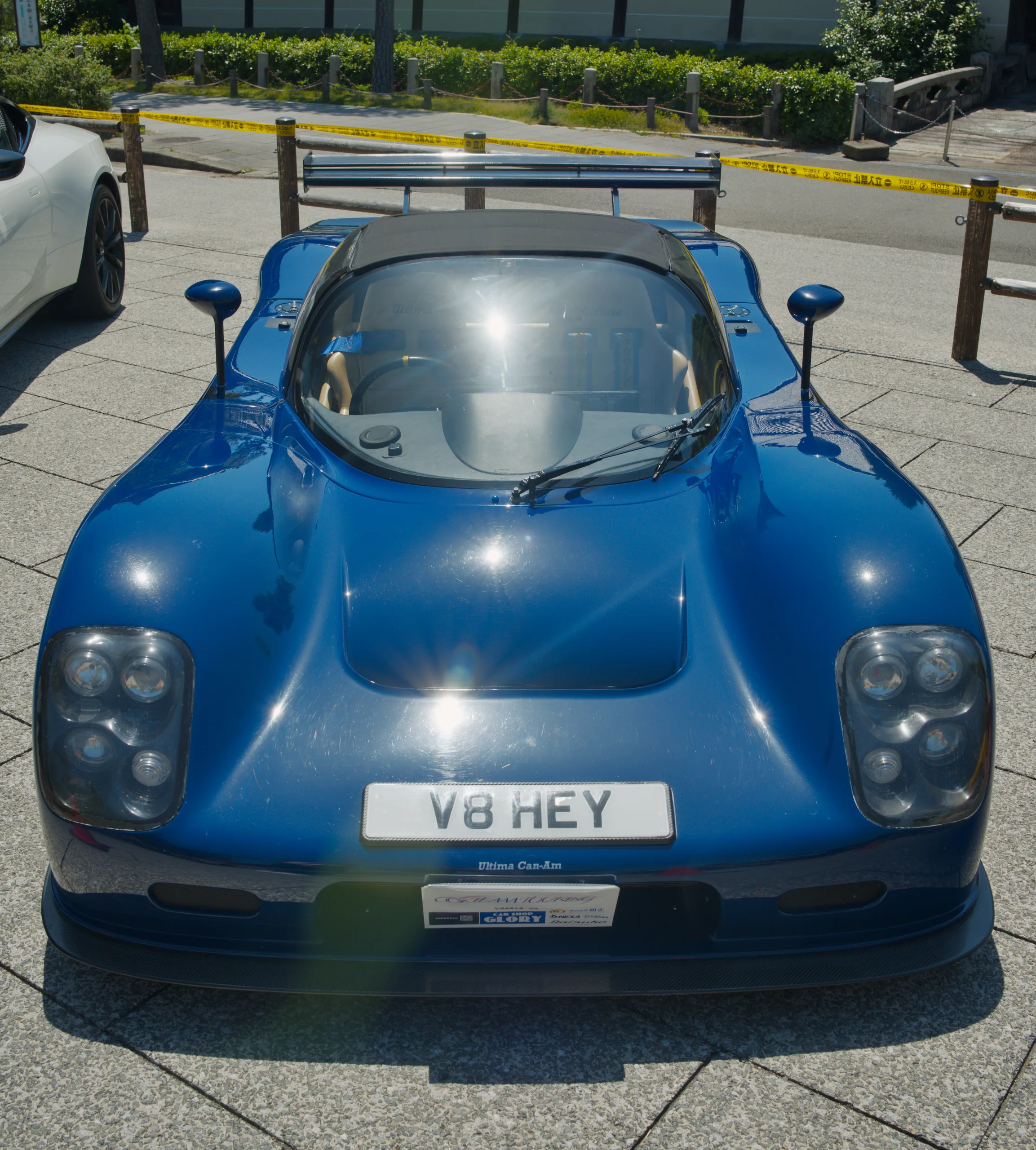 A blue sports car with a rounded, compact body at a car show with barriers and green foliage behind it.