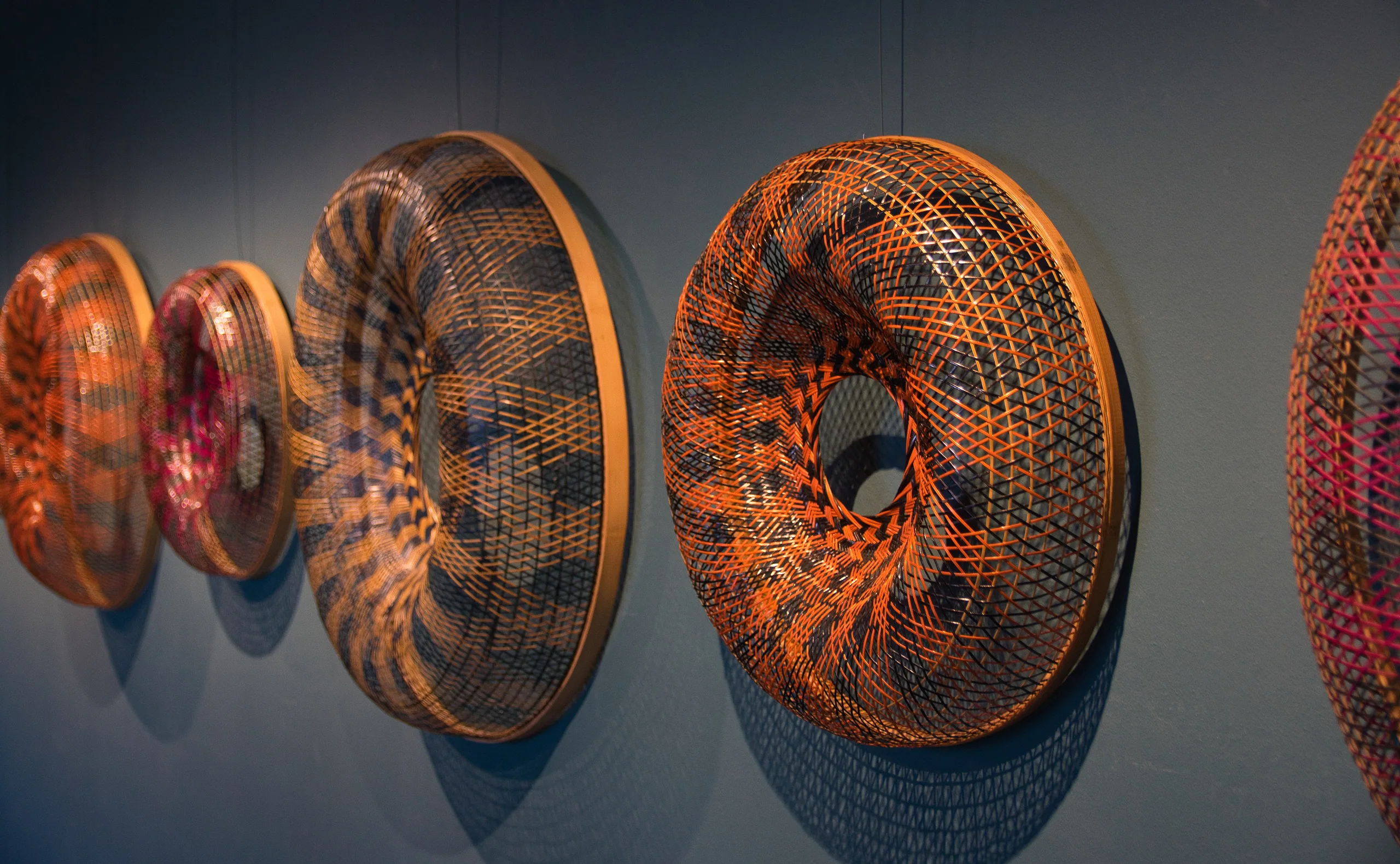 A row of large woven torus sculptures in orange and pink hung along a gallery wall, each one a spiral of fine cane.