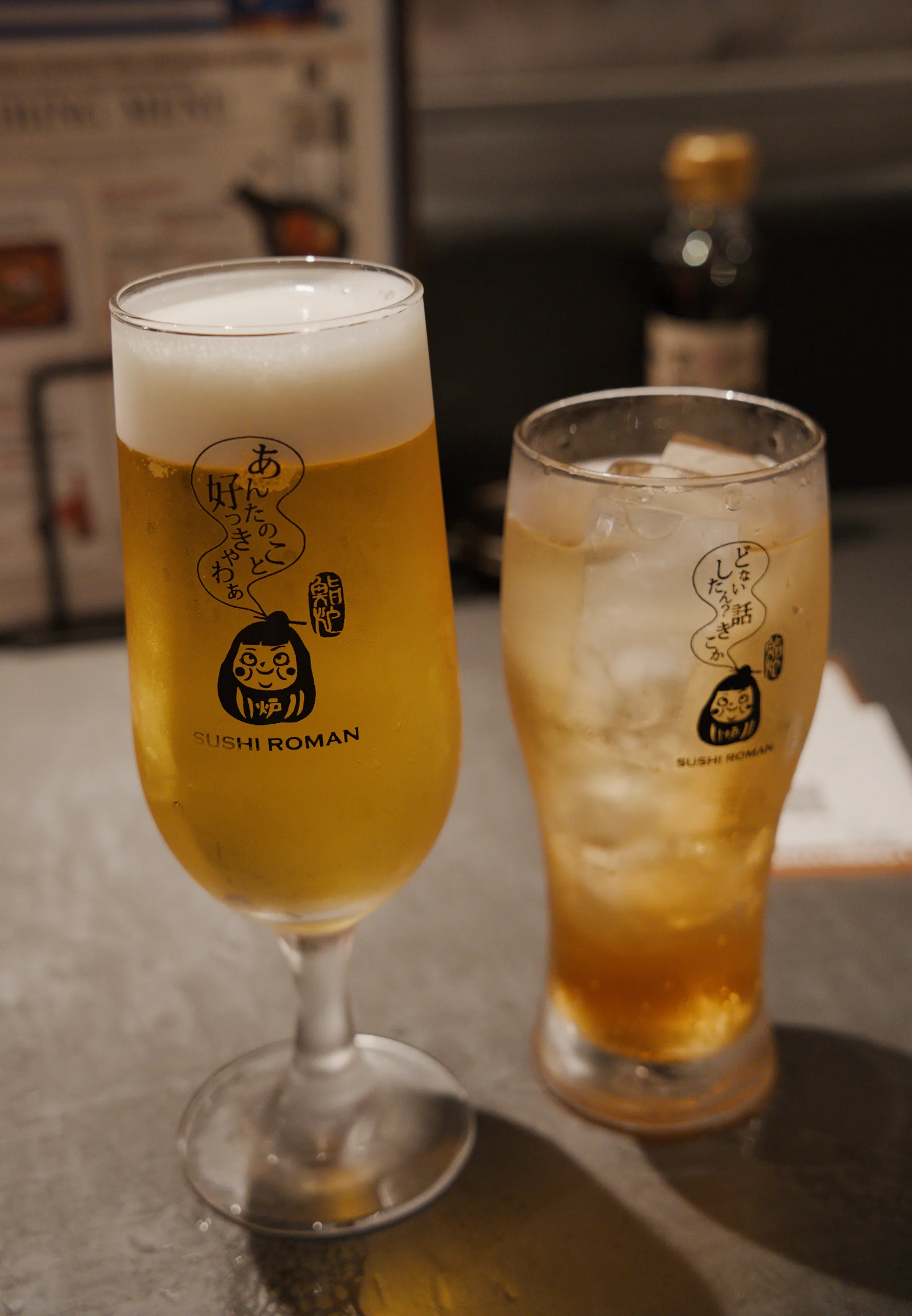 Two glasses of chilled beverages—one pale and one amber—on a table with restaurant branding.