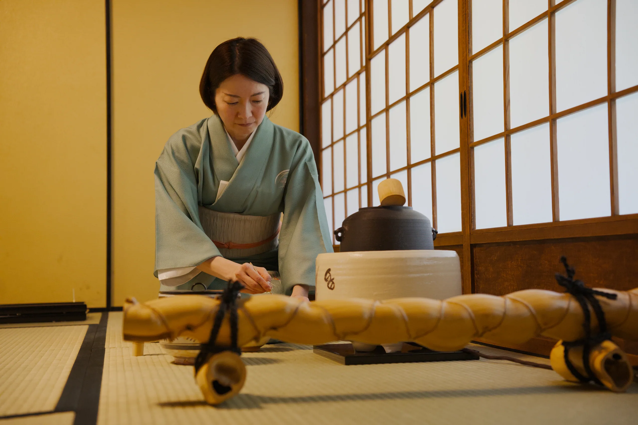Person in traditional green garment preparing tea at a low table in a Japanese room with latticed window screens.