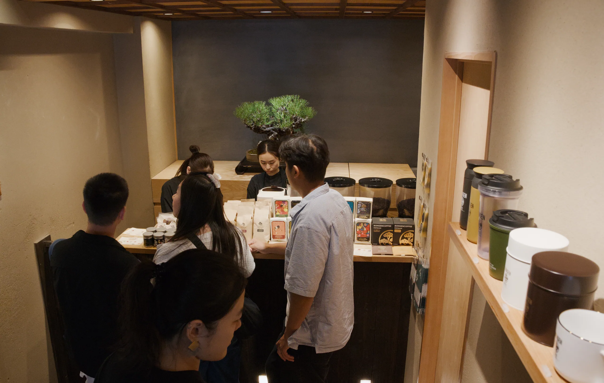 Customers at a counter in a small restaurant with warm lighting and a bonsai tree on the wall.