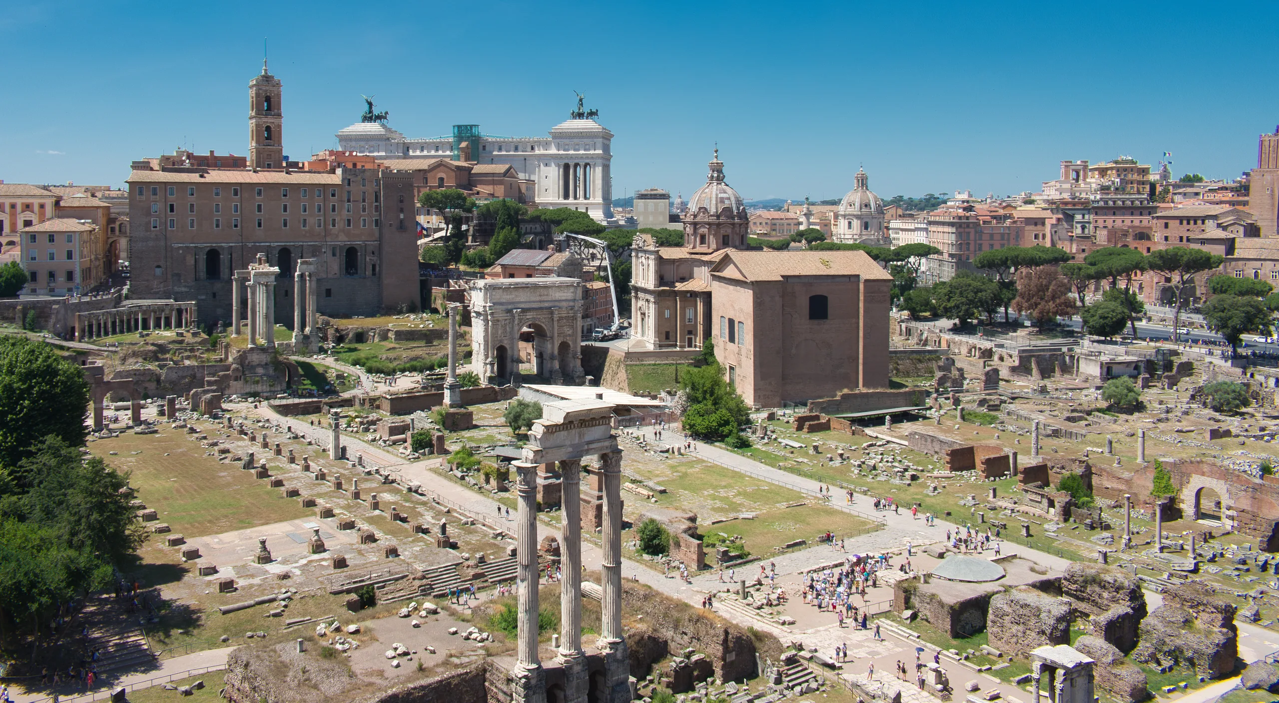 The Roman Forum seen along its length, standing columns and paved ruins with domes and towers of the city behind.